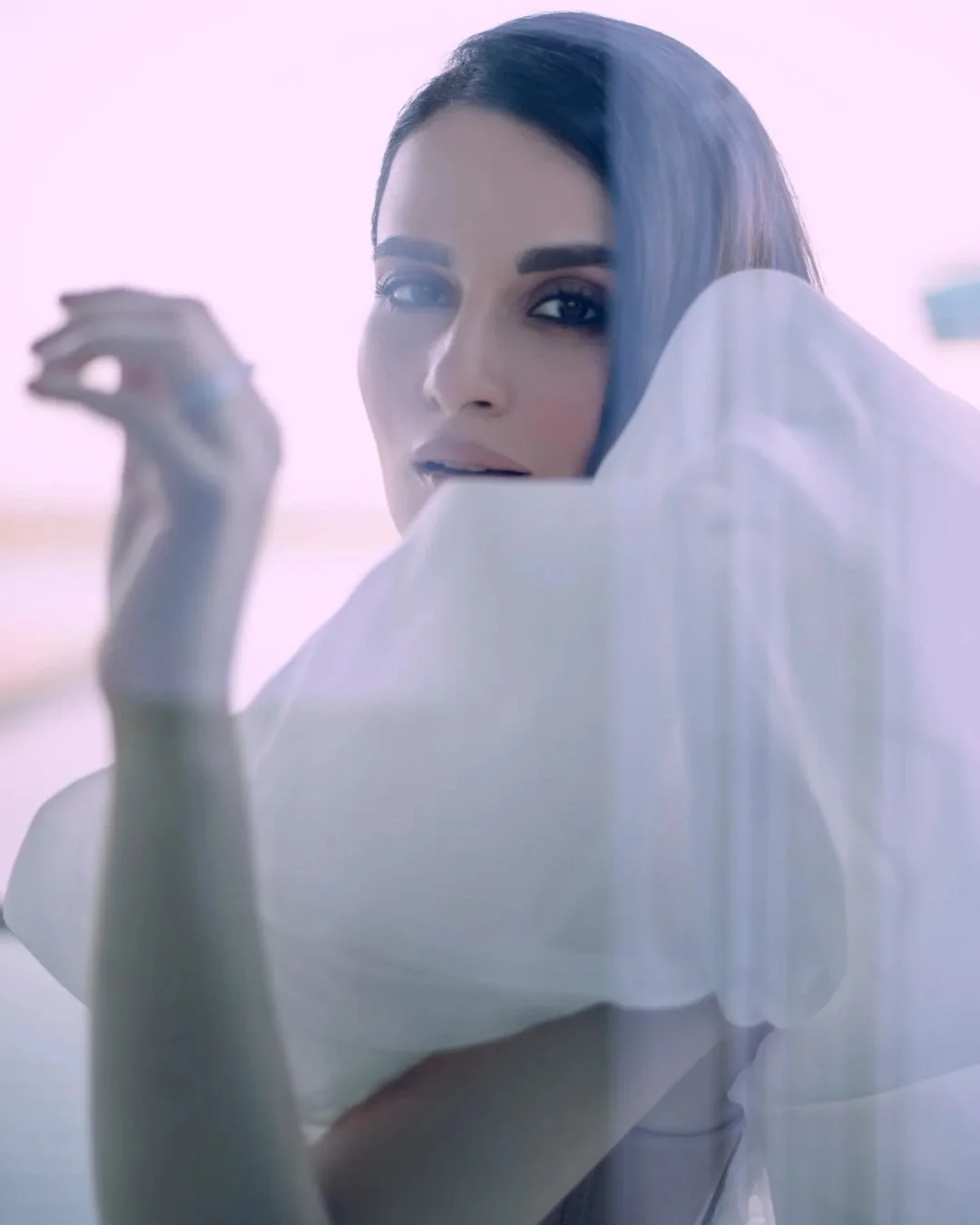 Close-up of a woman with dark hair and makeup, in a white shirt, looking at the camera with her hand near her face, in a brightly lit setting.