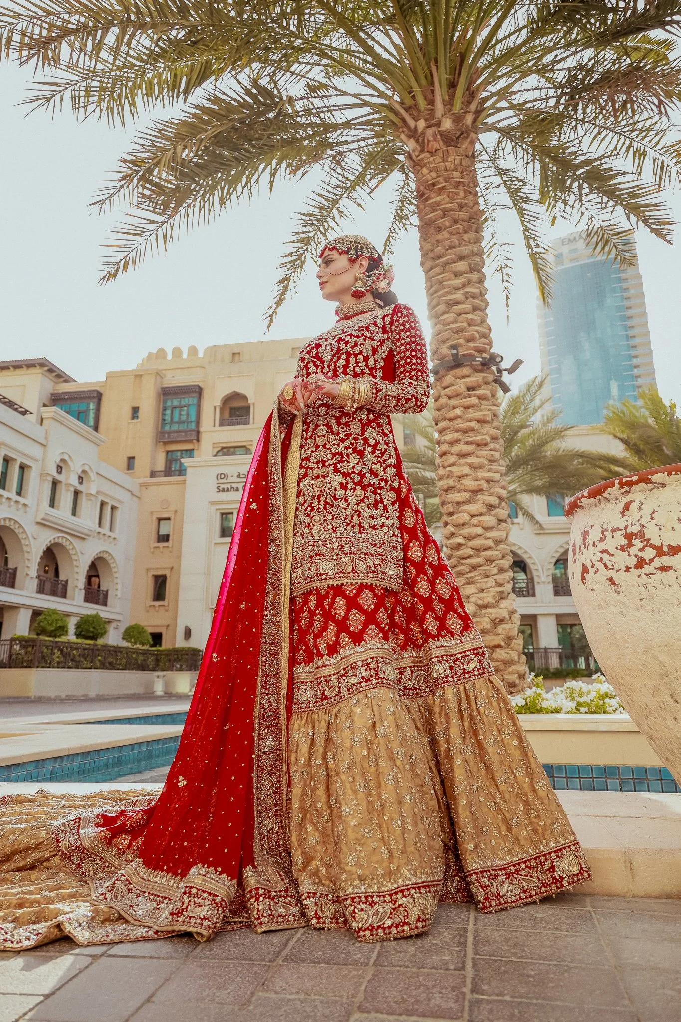 A woman in traditional Indian red and gold bridal attire standing next to a palm tree outdoors.