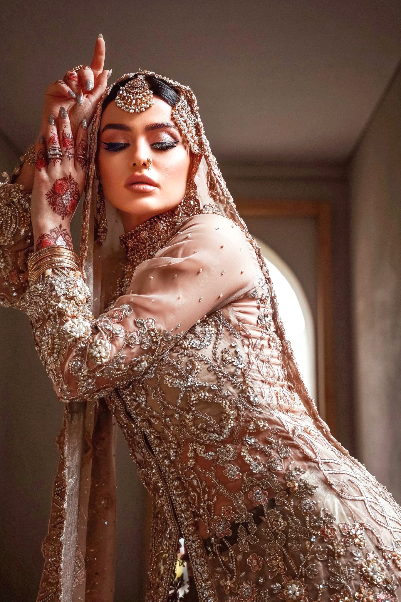 A woman in traditional intricate, heavily embroidered attire with jewelry and henna designs on her hands posing indoors.