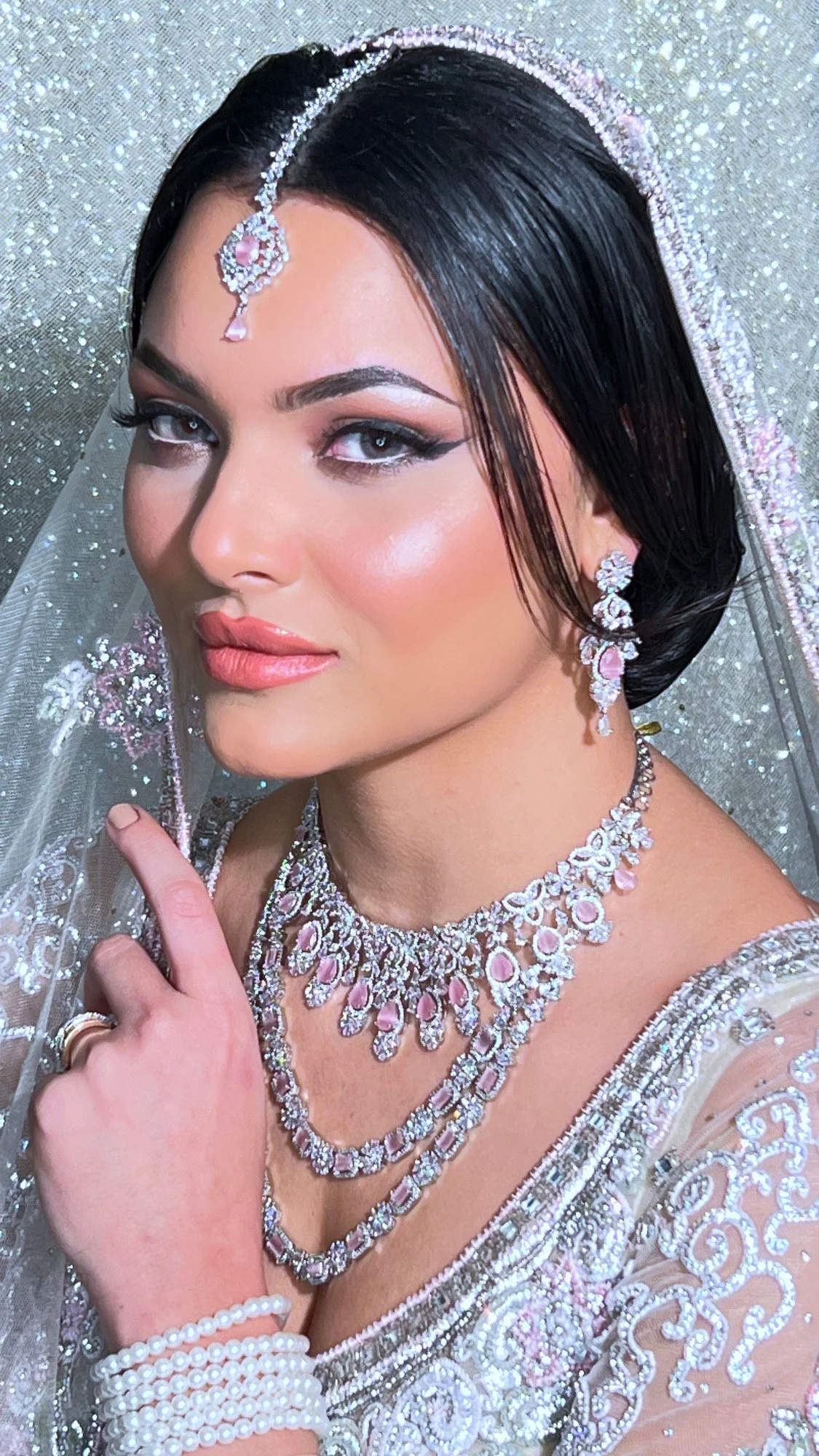 A woman dressed as a bride, wearing an ornate wedding dress and jewelry, with dark hair, earrings, a necklace, a tiara, and a veil with floral details, holding her hand near her face.