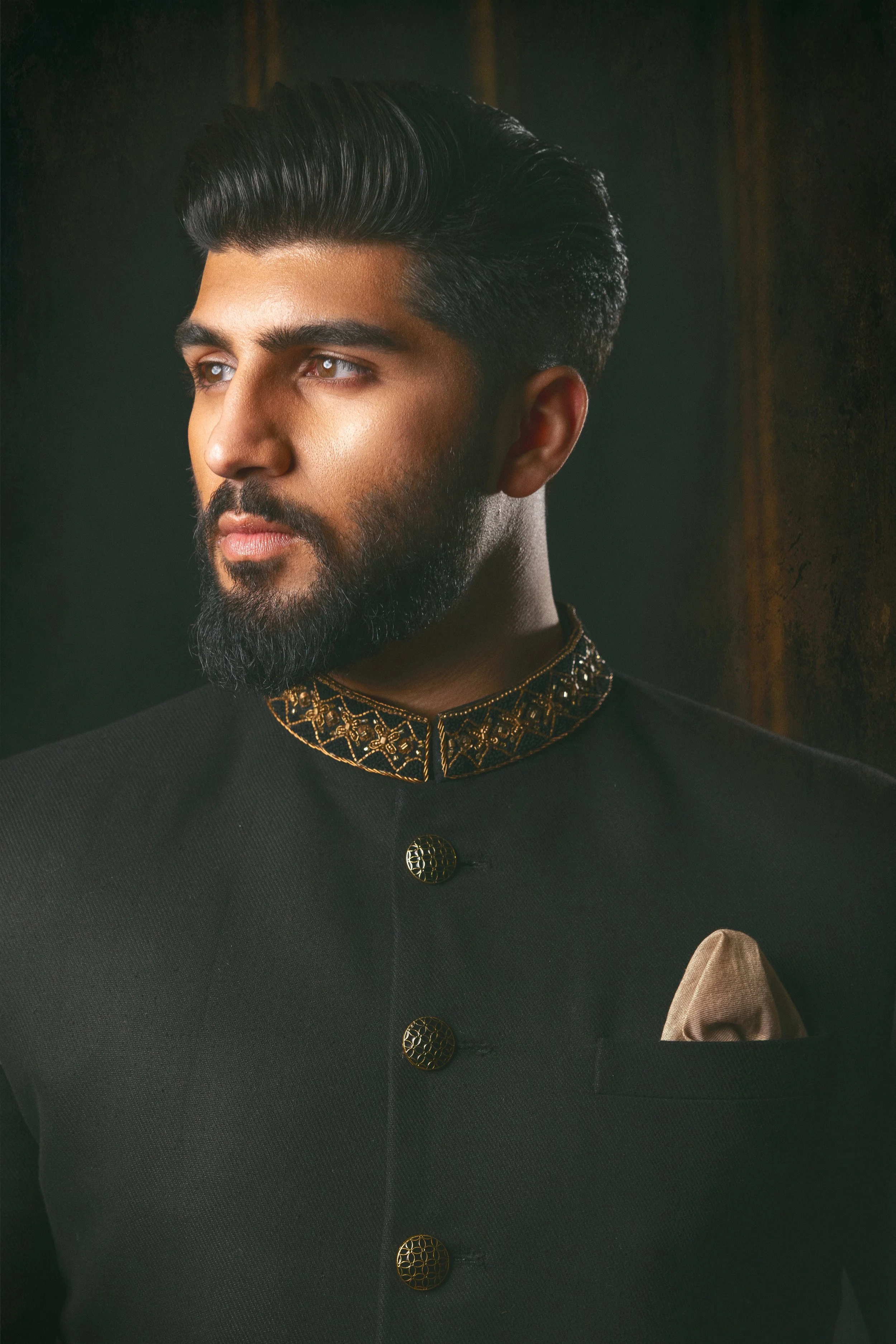 A man with a beard and styled hair wearing a black sherwani with gold embroidery on the collar and gold buttons, with a beige pocket square in the breast pocket
