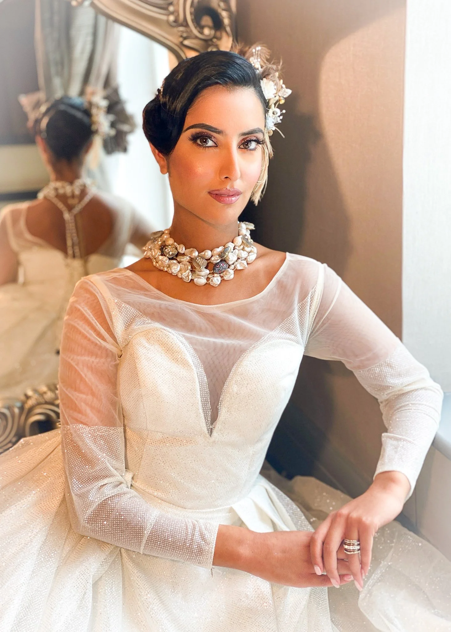 A woman dressed in a white wedding gown with sheer long sleeves, sitting near a mirror. She has dark hair styled in an elegant updo, adorned with a floral headpiece, and is wearing a pearl and floral necklace. Her makeup is sophisticated, and she is sitting with her hands folded, showing a wedding ring.