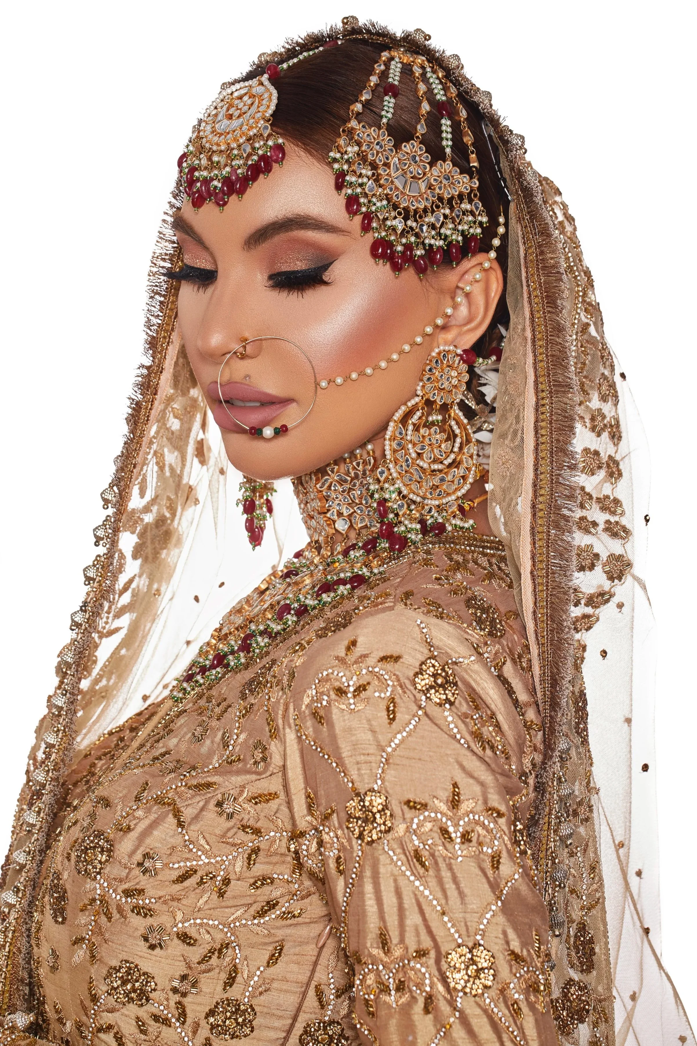 A woman wearing traditional South Asian bridal attire with intricate gold embroidery and jewelry, including a headpiece, earrings, nose ring, and necklace, with a veil and styled hair.
