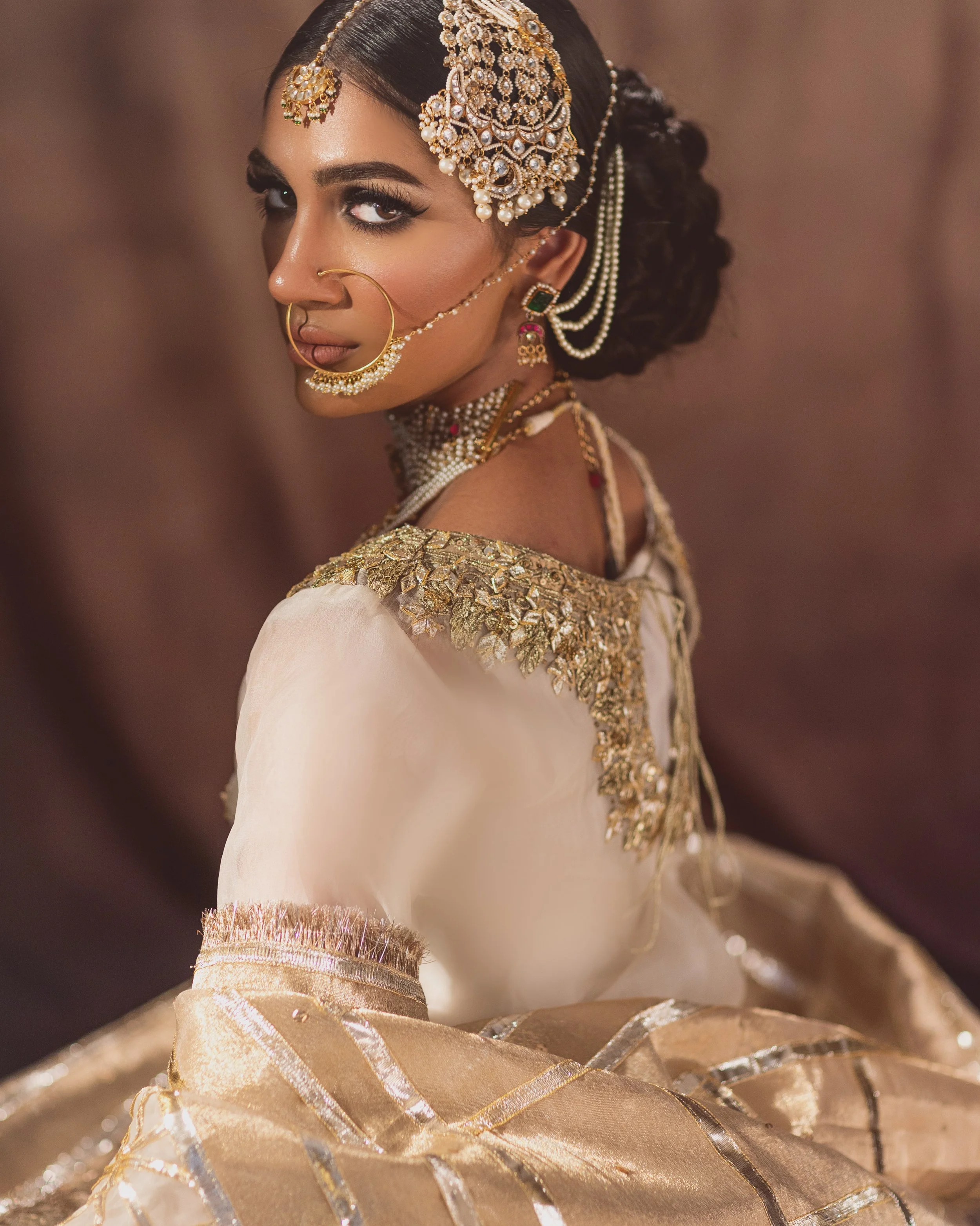 A woman dressed in traditional Indian bridal attire with intricate gold jewelry, including a headpiece, earrings, nose ring, and layered necklaces, posing with her head turned slightly over her shoulder.