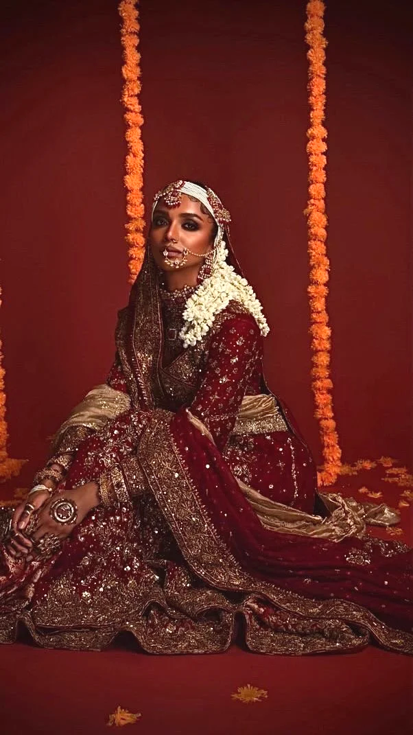 A woman dressed in traditional Indian bridal attire, sitting on the ground against a red background decorated with marigold flower garlands.