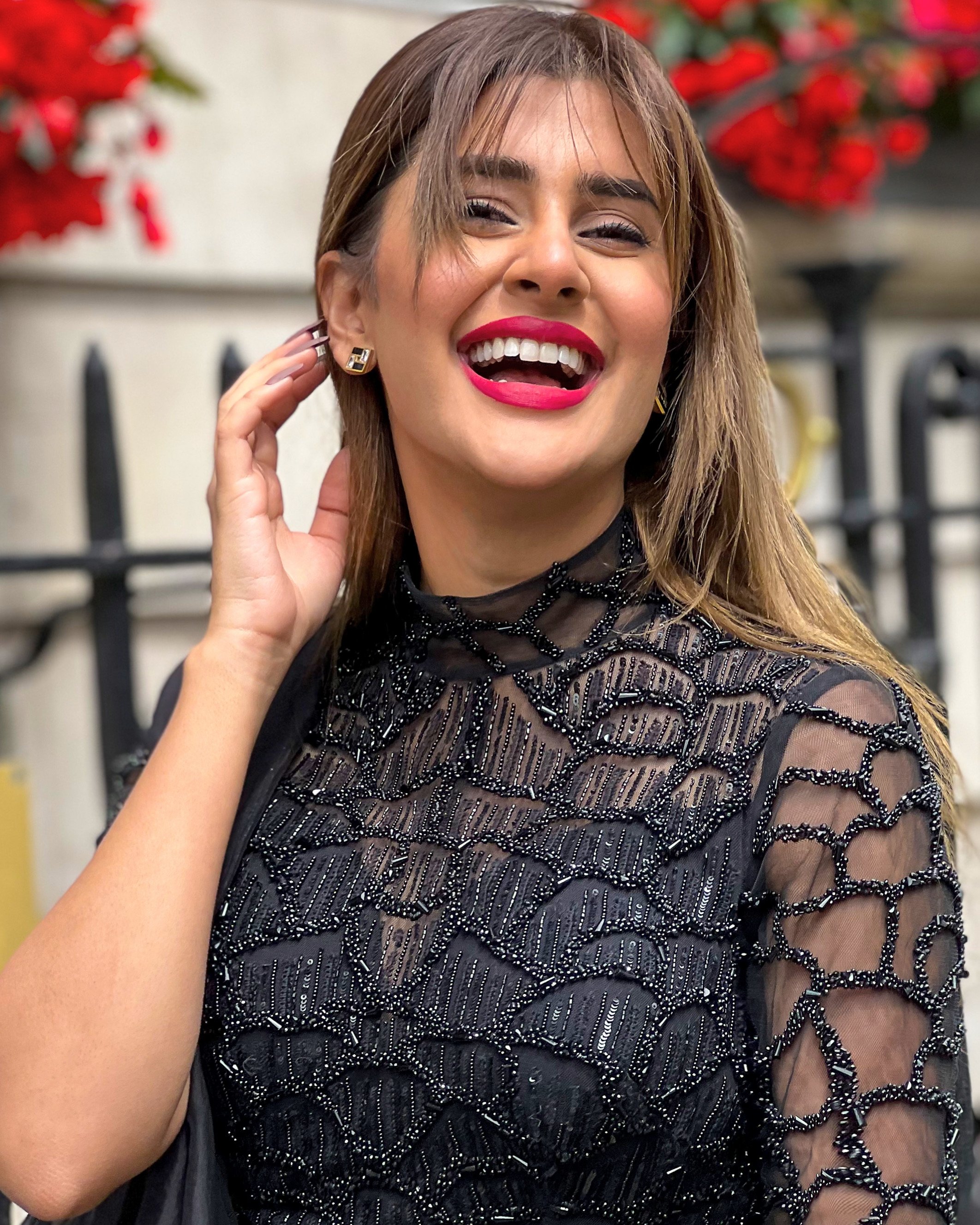 A woman with brown hair, bright pink lipstick, and a black beaded dress, smiling and touching her ear.