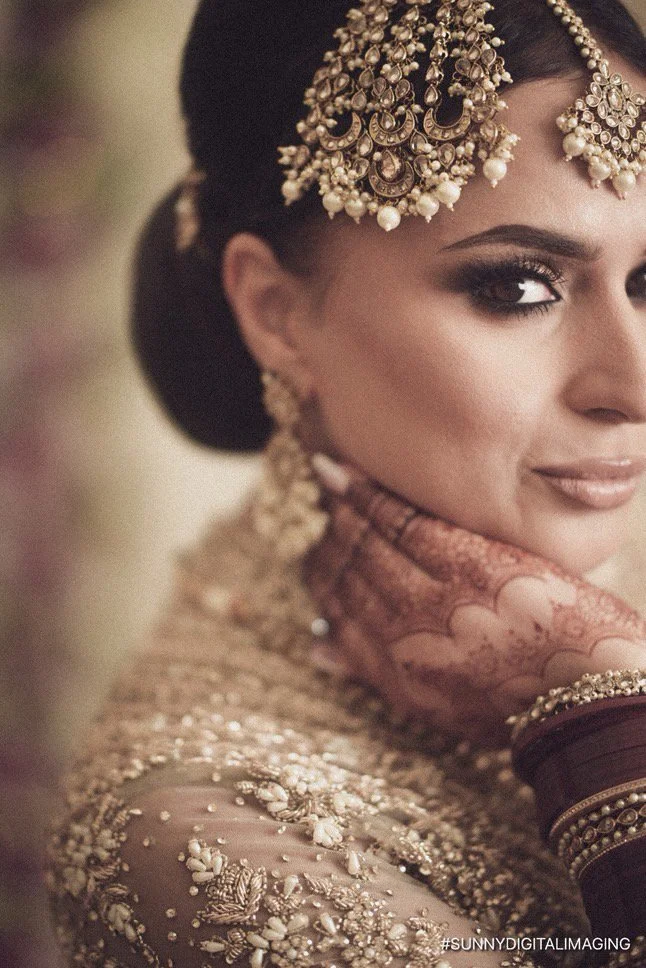 Close-up of a woman dressed in traditional Indian attire, wearing intricate jewelry including a maang tikka, earrings, and bangles, with her hair styled in an updo.