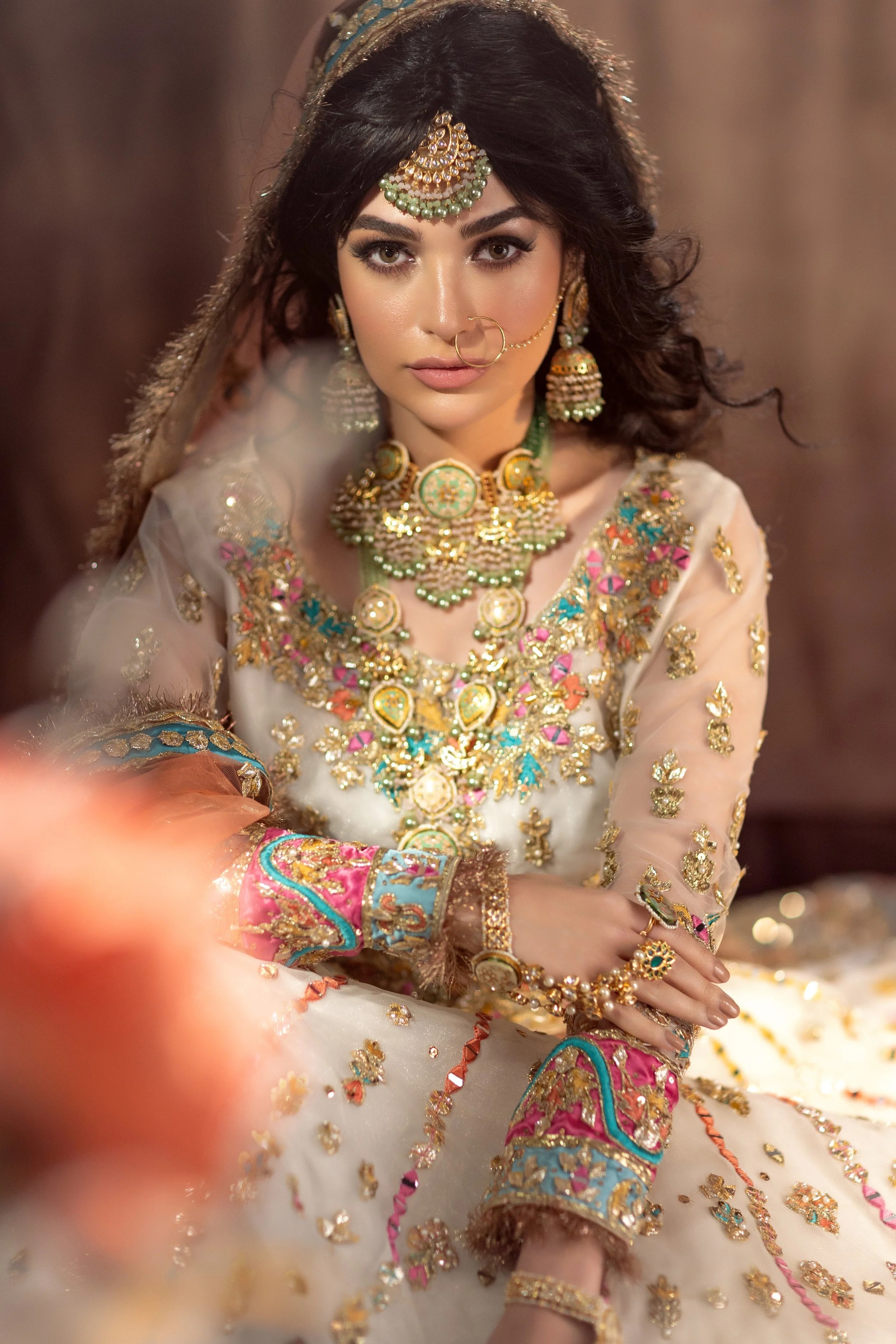 A woman wearing traditional Indian bridal attire decorated with gold jewelry, including multiple necklaces, earrings, a maang tikka, bangles, and a nose ring, is captured in a close-up portrait.