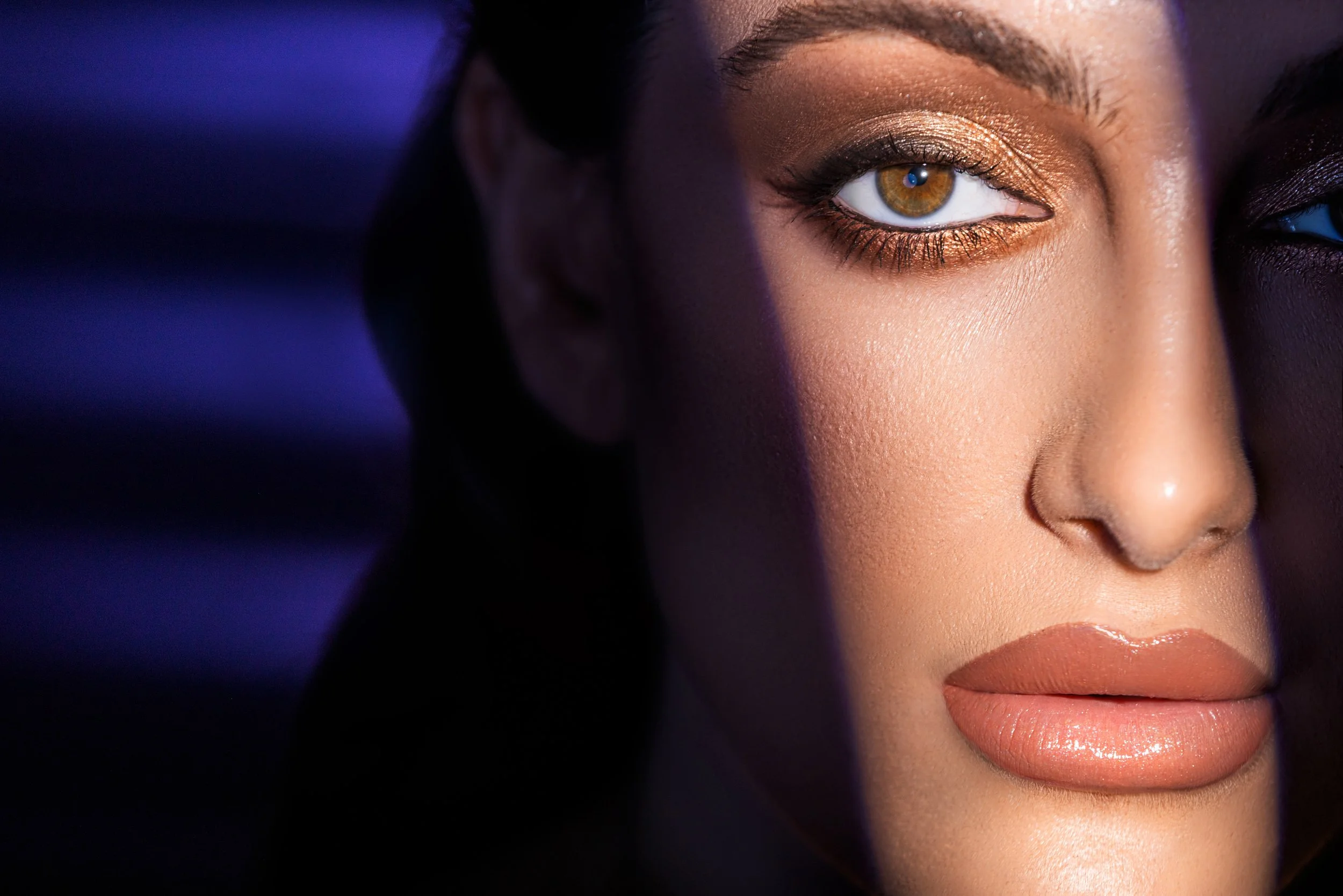 Close-up of a woman's face with makeup, showing her brown eye with golden eyeshadow and glossy lips, with a shadow partially covering her face.