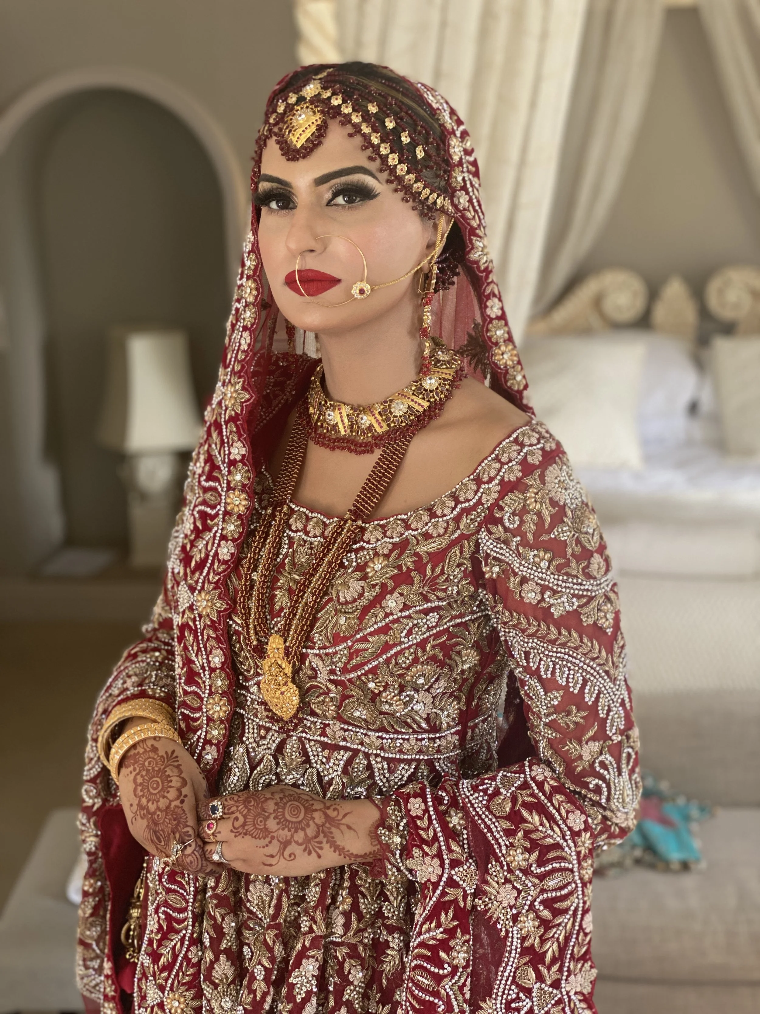 A woman dressed in traditional South Asian bridal attire, wearing a heavily embellished red and gold embroidered outfit with matching jewelry, including a head covering, necklaces, earrings, nose ring, and henna designs on her hands.