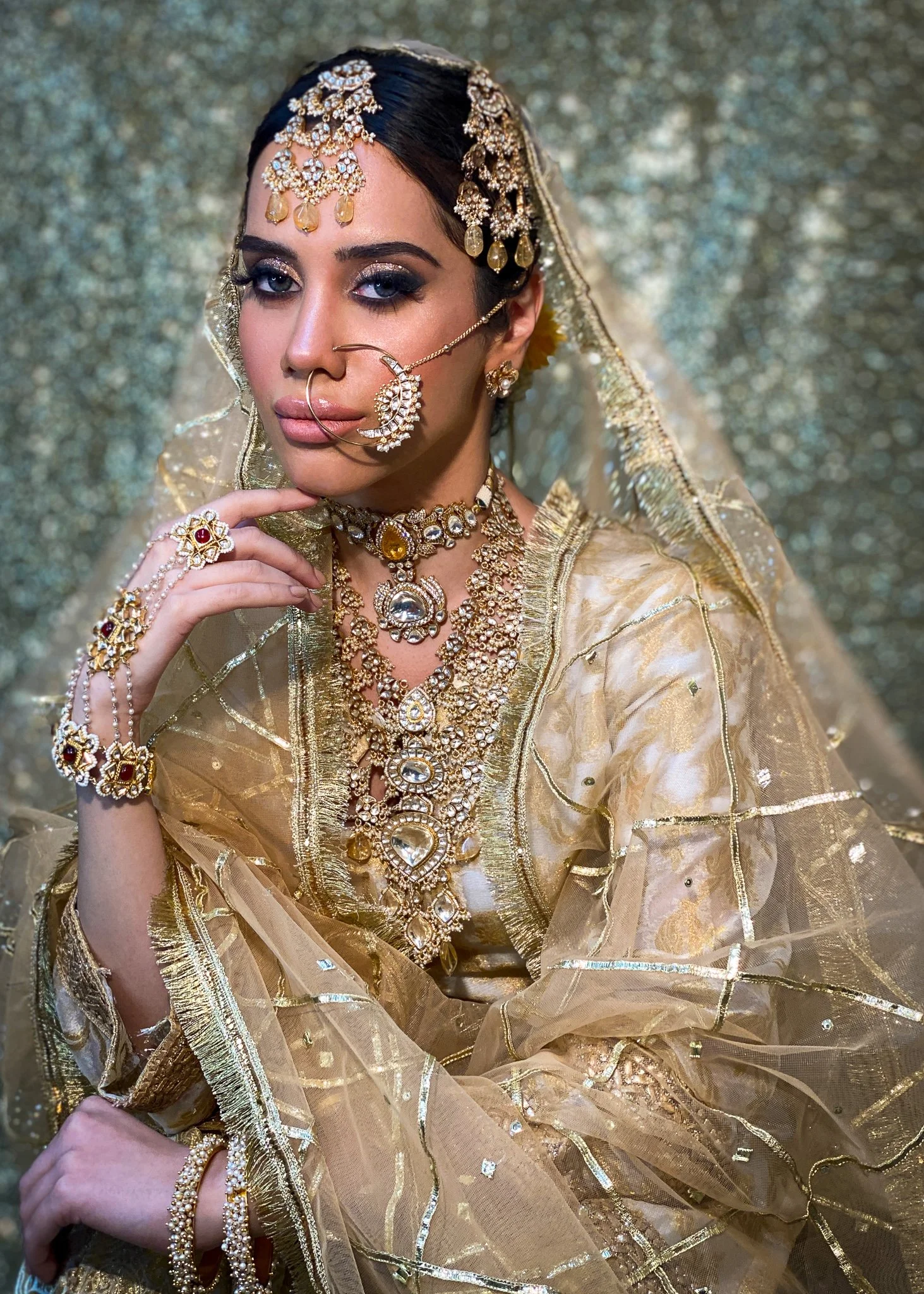A woman dressed in traditional Indian bridal attire, wearing elaborate gold jewelry including a headpiece, nose ring, necklaces, bracelets, and rings, with a gold embroidered outfit and veil, against a glittering gold background.