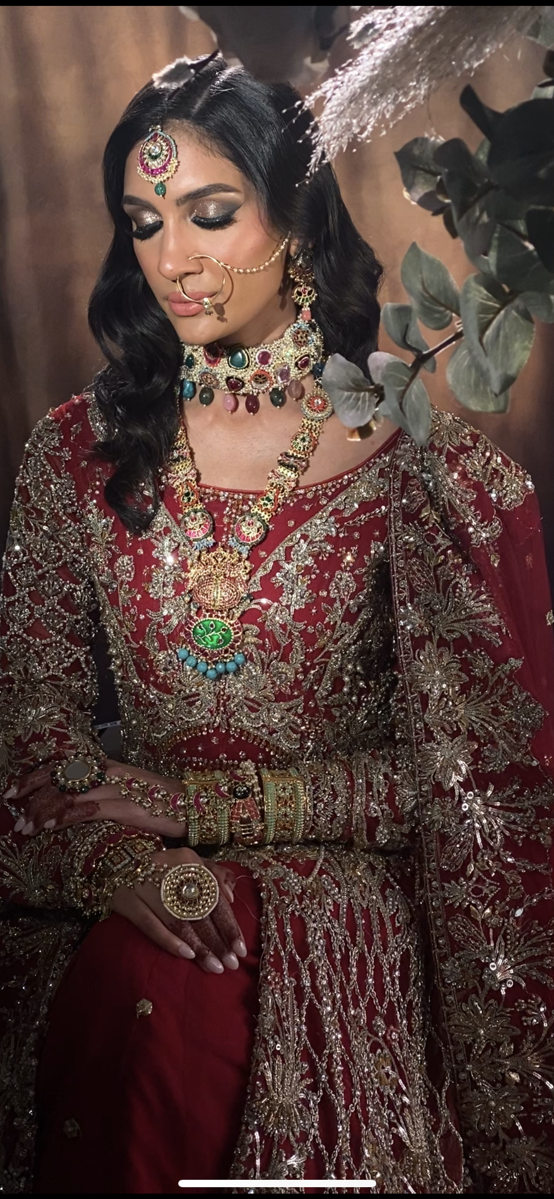 A woman dressed in traditional Indian bridal attire with elaborate jewelry and embroidery, standing in front of a floral arrangement.
