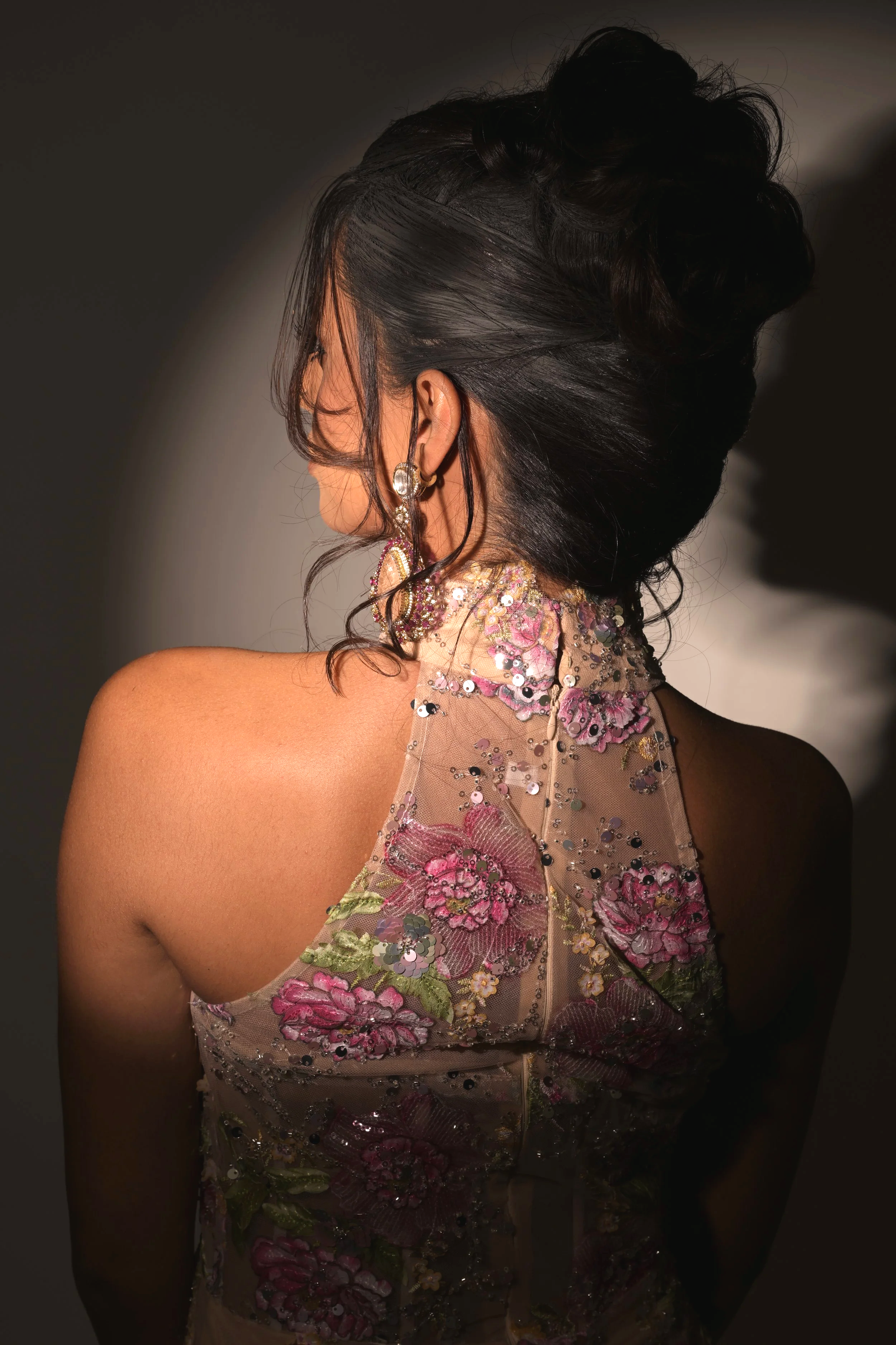 A woman with dark hair styled in an elegant updo, wearing large earrings and a sheer, embroidered dress with floral patterns and sequins, standing against a dark background.