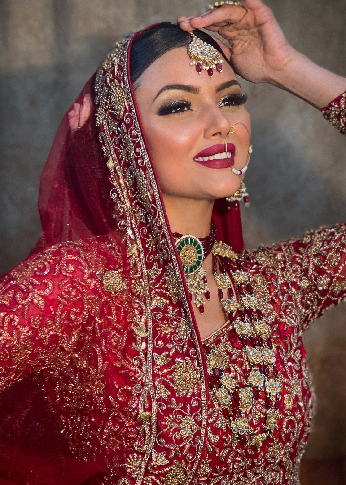 A woman dressed in traditional South Asian bridal attire, wearing a richly embroidered red and gold bridal dress, elaborate jewelry, and a decorative veil, posing with a smile.