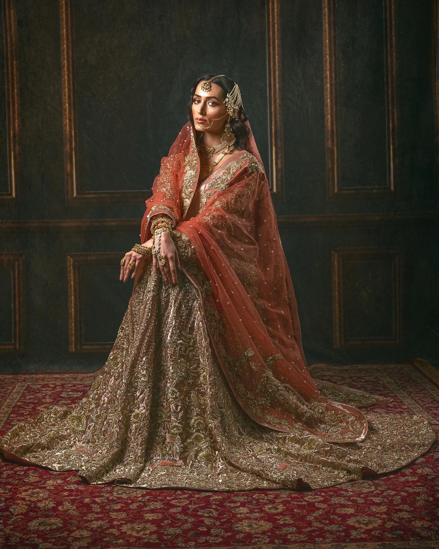 A woman dressed in elaborate traditional Indian wedding attire, including a gold and red embroidered lehenga, dupatta, and intricate jewelry, sitting on a richly patterned carpet in a dark, elegant room with wood-paneled walls.