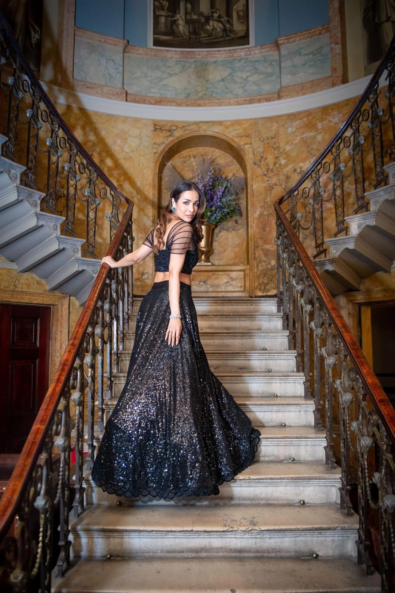 A woman in a black, glittery evening gown standing on a grand staircase inside an ornate building.