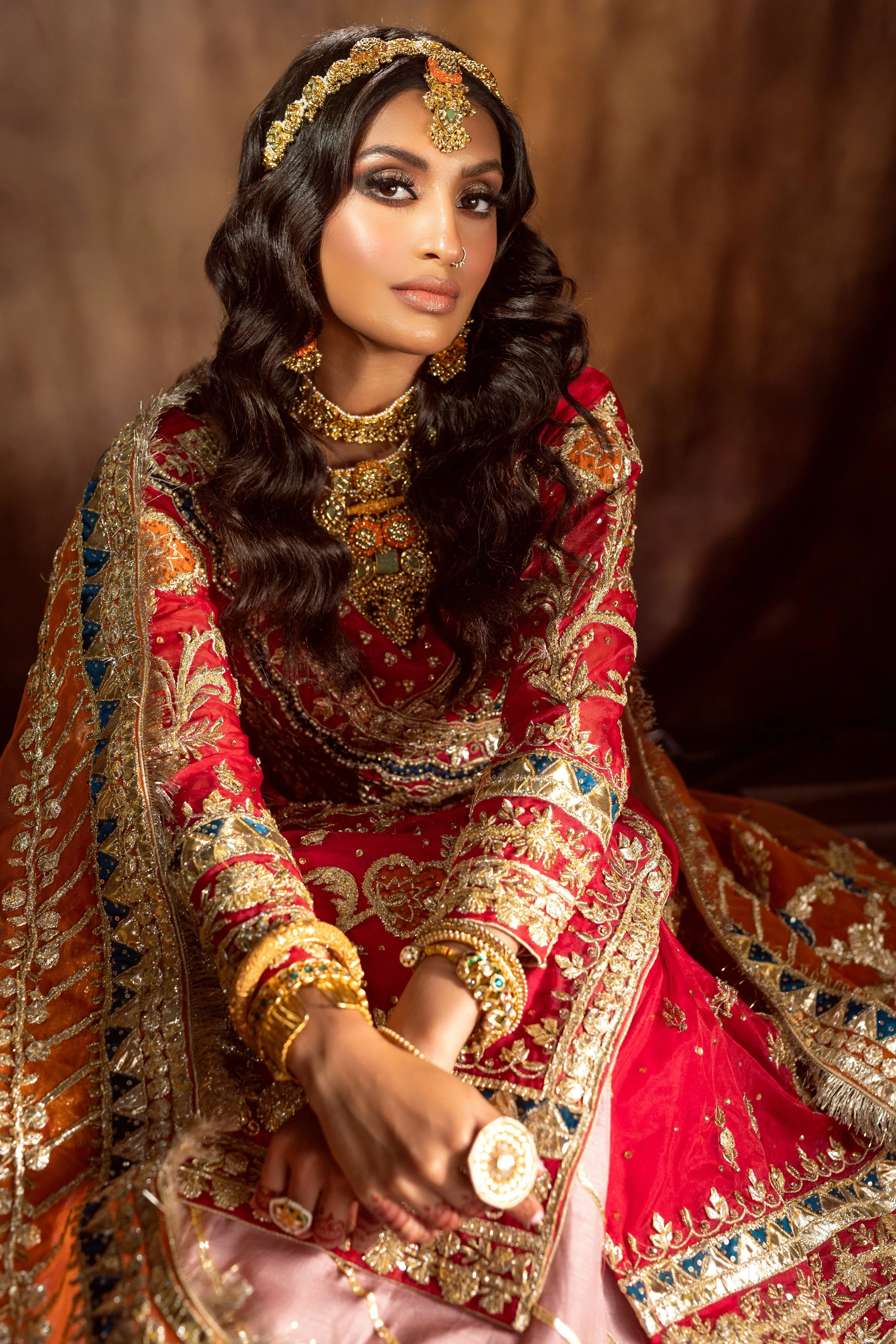 A woman dressed in traditional South Asian bridal attire with intricate gold embroidery, wearing heavy jewelry including a headpiece, earrings, necklace, bangles, and rings, seated against a brown background.