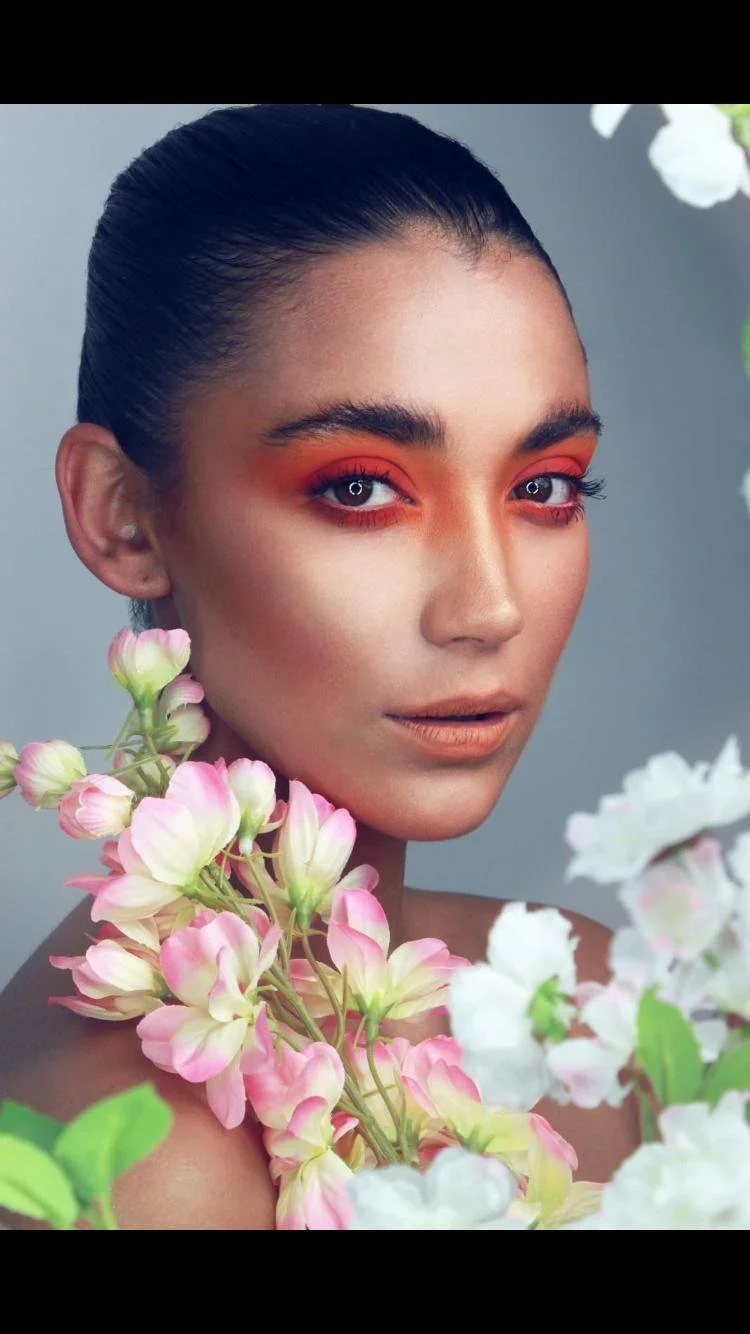 A woman with dark hair tied back, wearing orange eye makeup, is surrounded by pink and white flowers.