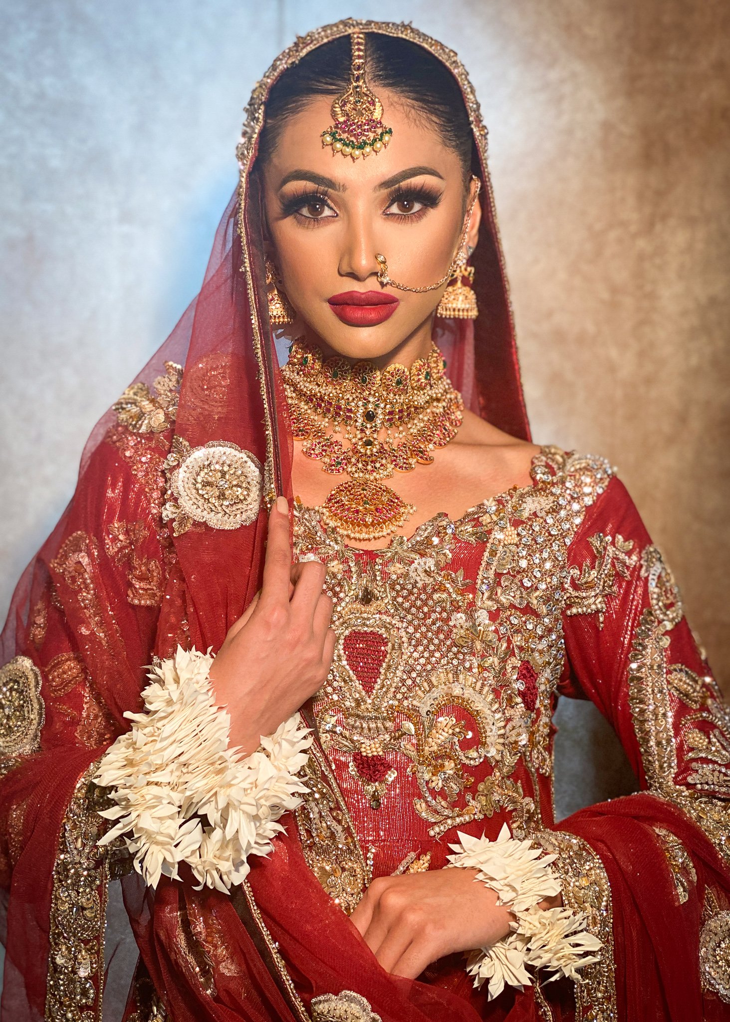 A woman dressed in traditional, heavily embroidered red bridal attire with gold and silver embellishments, wearing intricate jewelry including a headpiece, earrings, necklace, nose ring, and floral bracelets, against a neutral background.