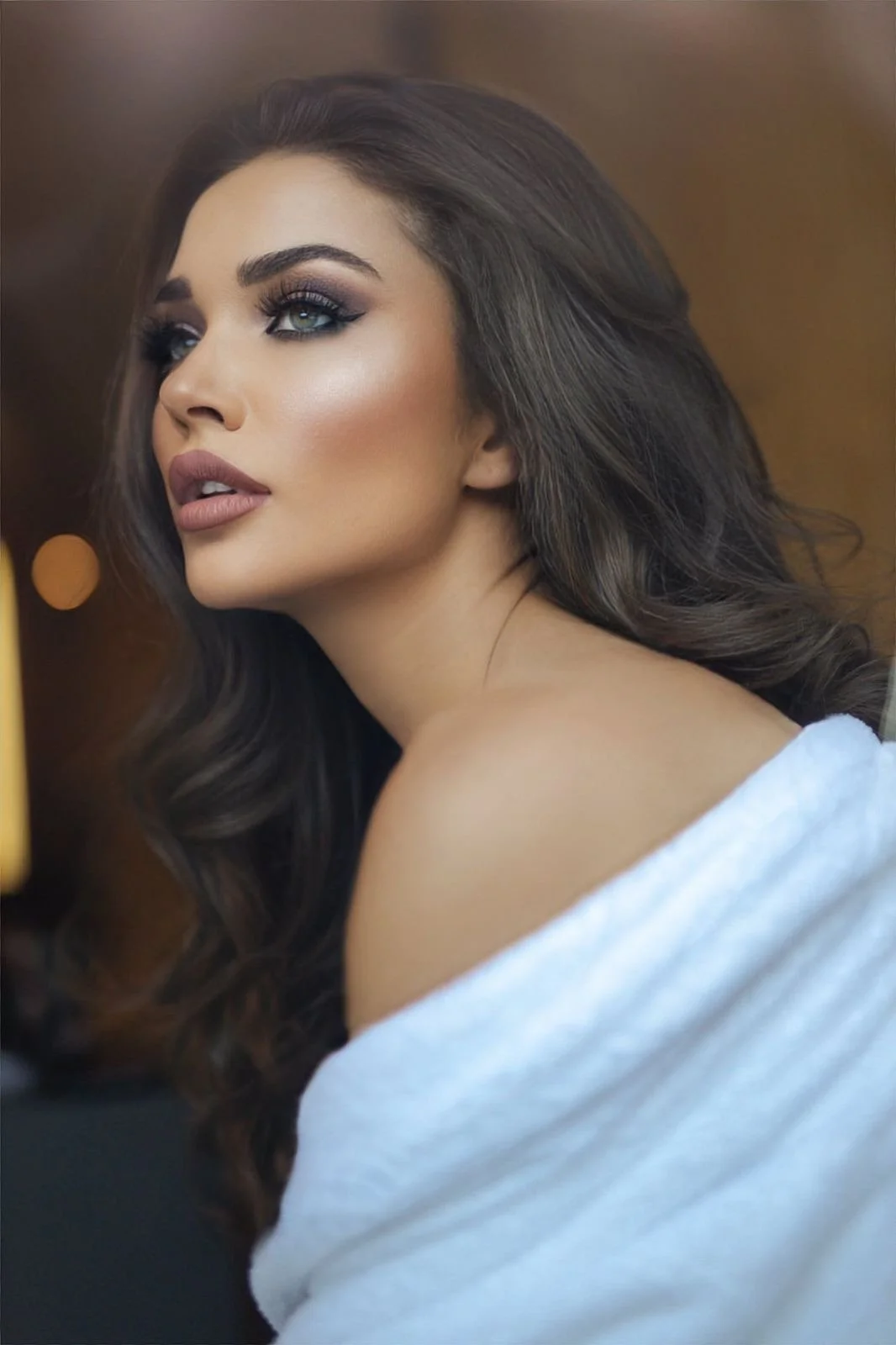 A woman with long, wavy brown hair and makeup, wearing a white towel draped over her shoulder.