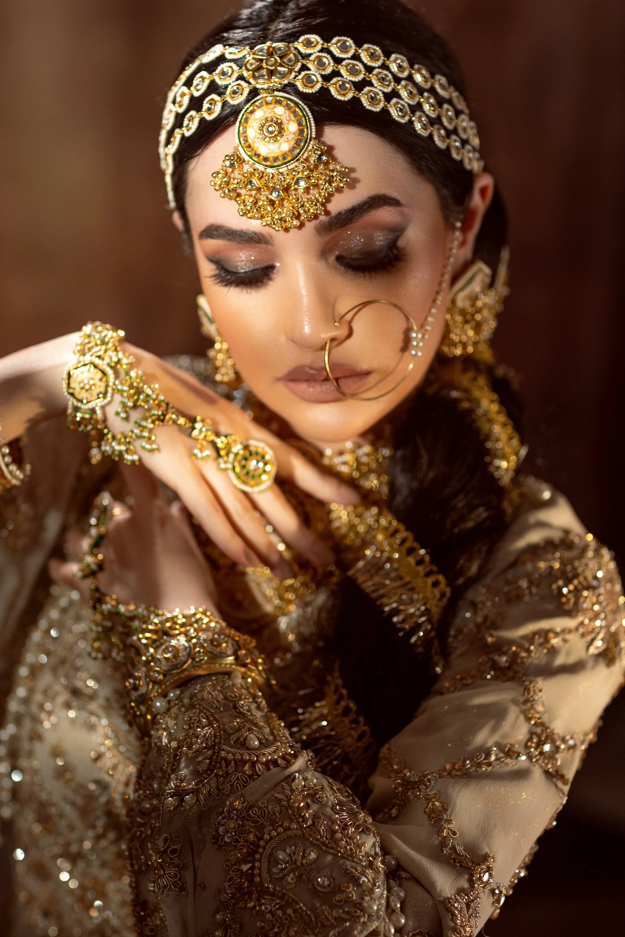 A woman dressed in traditional Indian jewelry and attire, with elaborate gold jewelry including a headpiece, earrings, nose ring, and bracelets, posing with her eyes closed and her hands near her face.