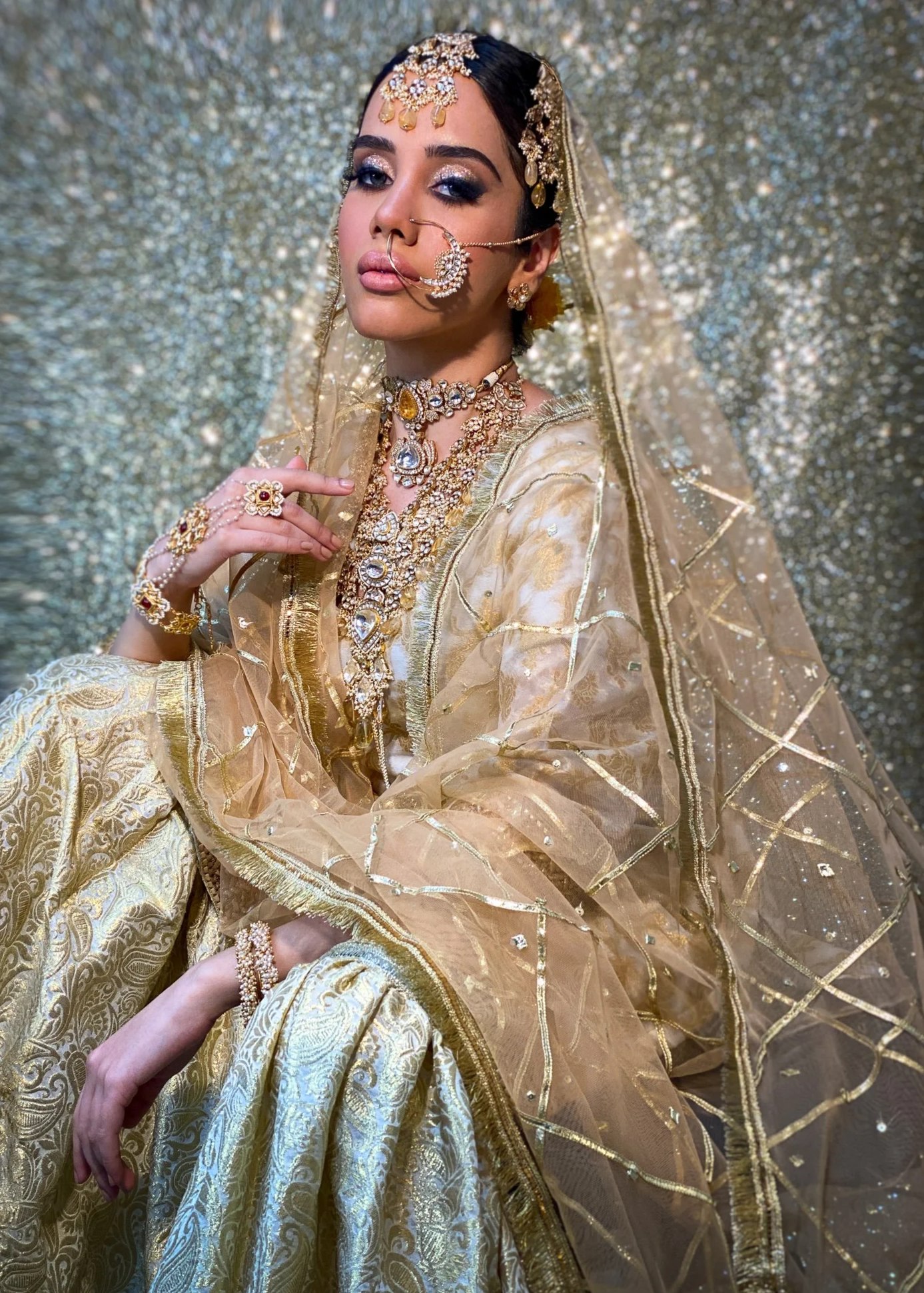 A woman dressed in traditional Indian bridal attire with intricate gold jewelry, a sheer gold embroidered dupatta, and matching golden embroidered clothing. She is posing against a glittery background, with jewelry including a headpiece, nose ring, necklace, rings, and bangles.