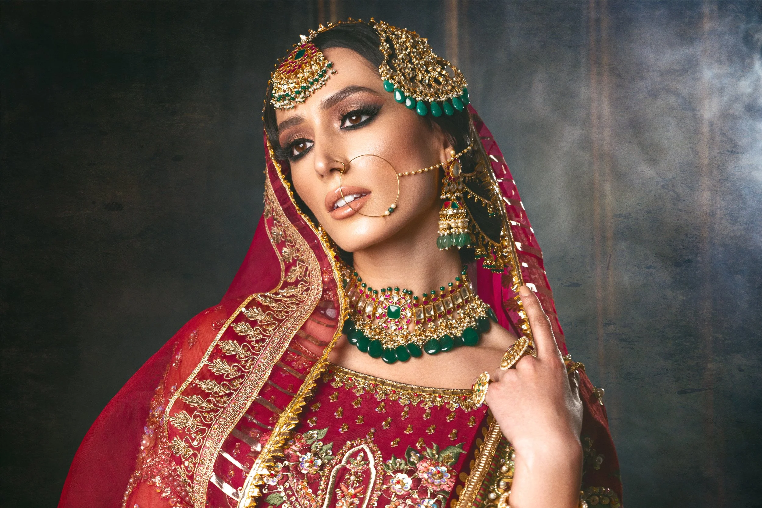 A woman dressed in traditional Indian bridal attire with elaborate jewelry, including a red and gold embroidery dress, a headpiece, earrings, necklace, nose ring, and rings, posing against a dark background.