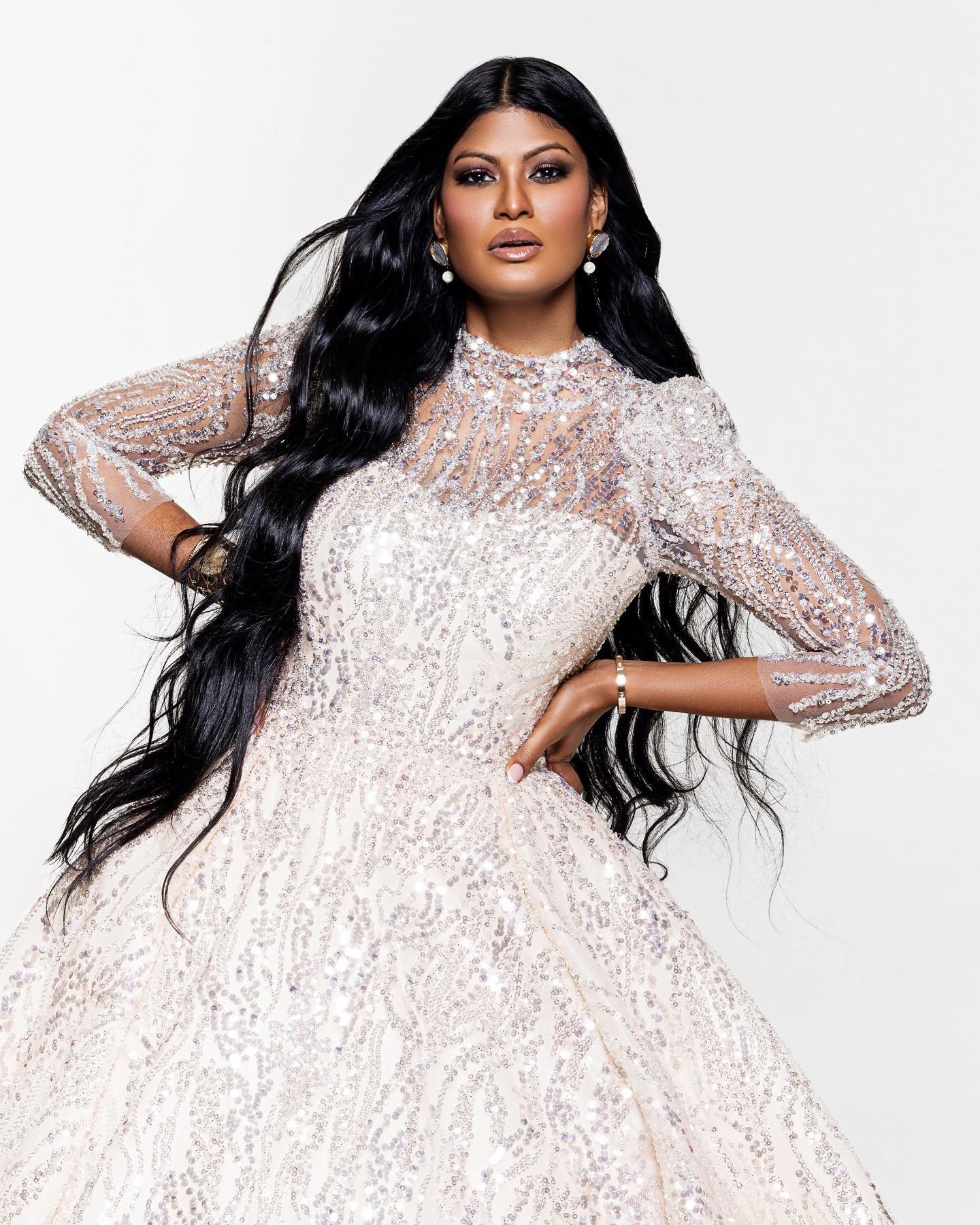 A woman with long black hair wearing a sparkling, intricate off-white gown and gold jewelry, posing with one hand on her hip against a plain background.