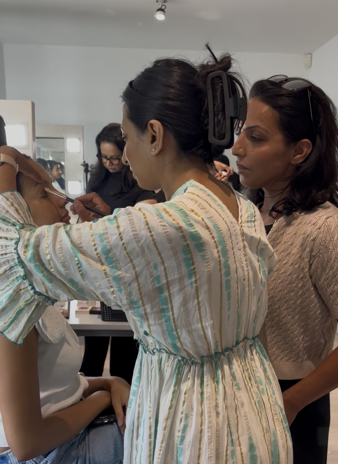 A woman is applying makeup to another woman’s face while others observe in a makeup studio or classroom setting.