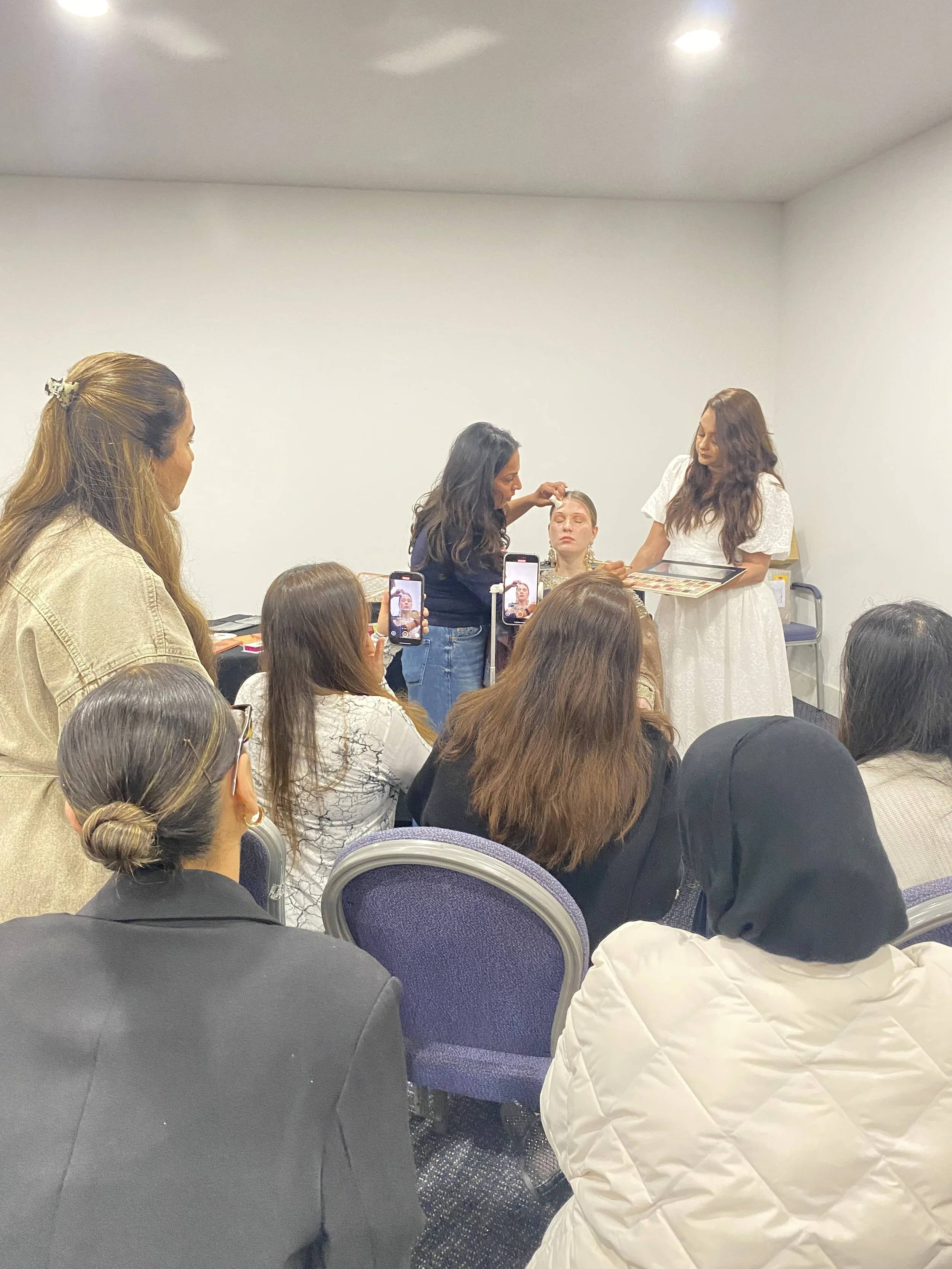 Women participating in a makeup demonstration in a room with white walls, with several attendees taking photos or videos with their phones.