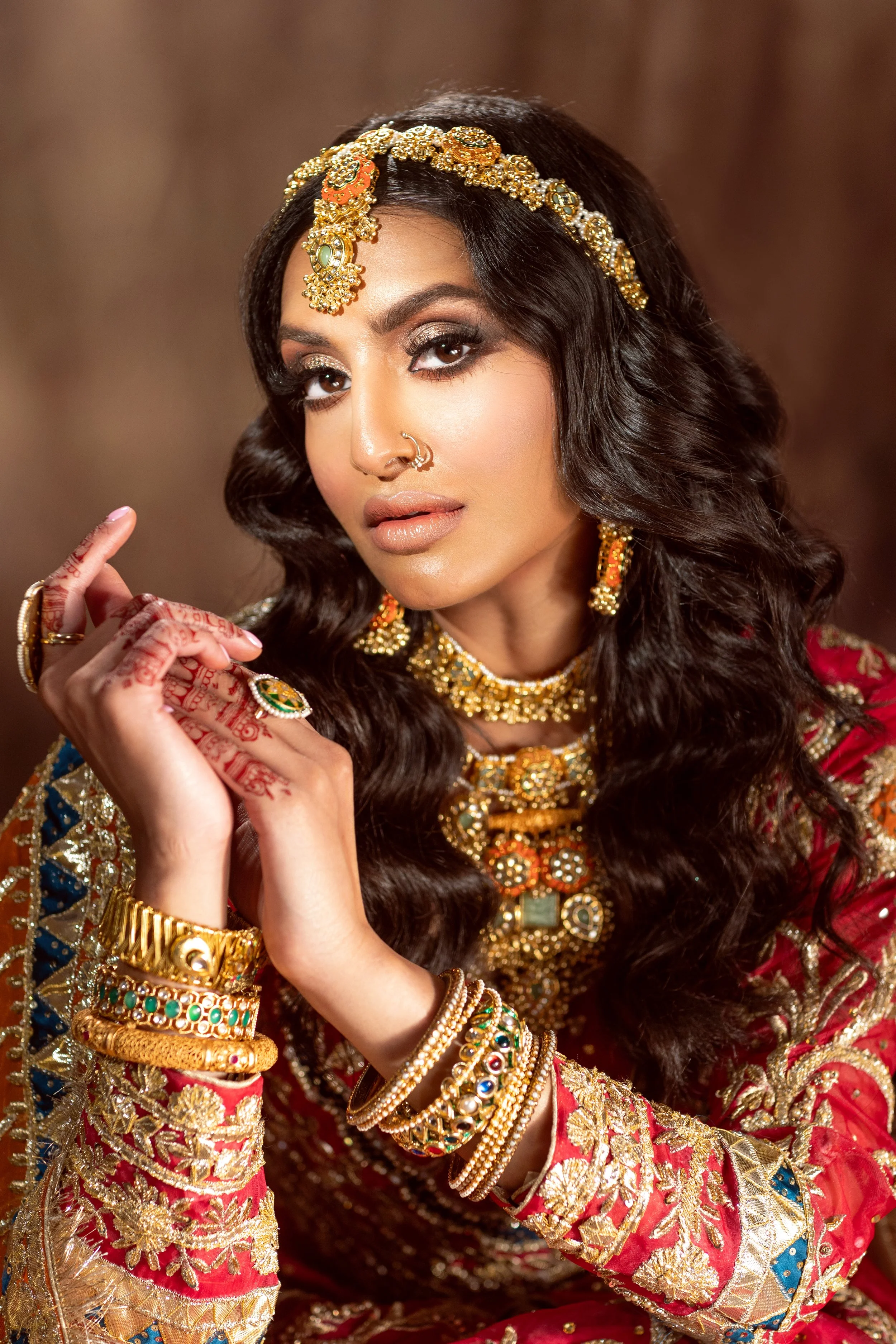 A woman dressed in traditional Indian attire, adorned with elaborate gold jewelry and intricate henna on her hands, against a warm background.