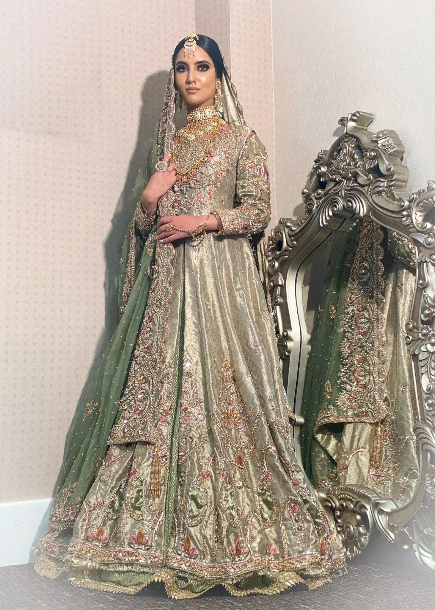 Woman dressed in a traditional, heavily embroidered bridal outfit with jewelry, standing next to an ornate mirror.