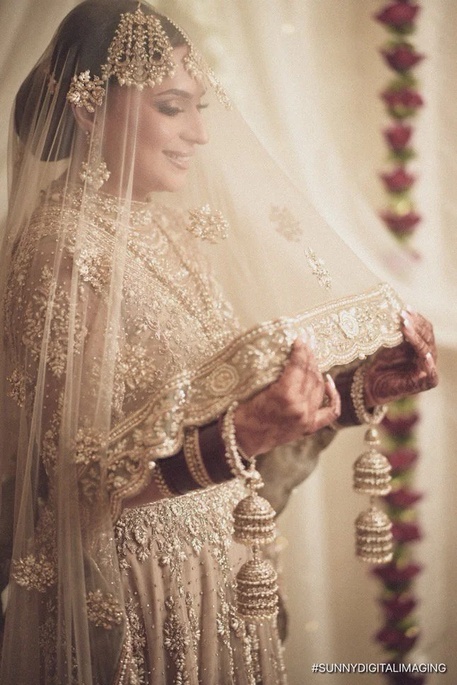 A bride wearing a heavily embroidered beige and gold wedding dress with traditional jewelry, holding a dupatta or veil, smiling softly.