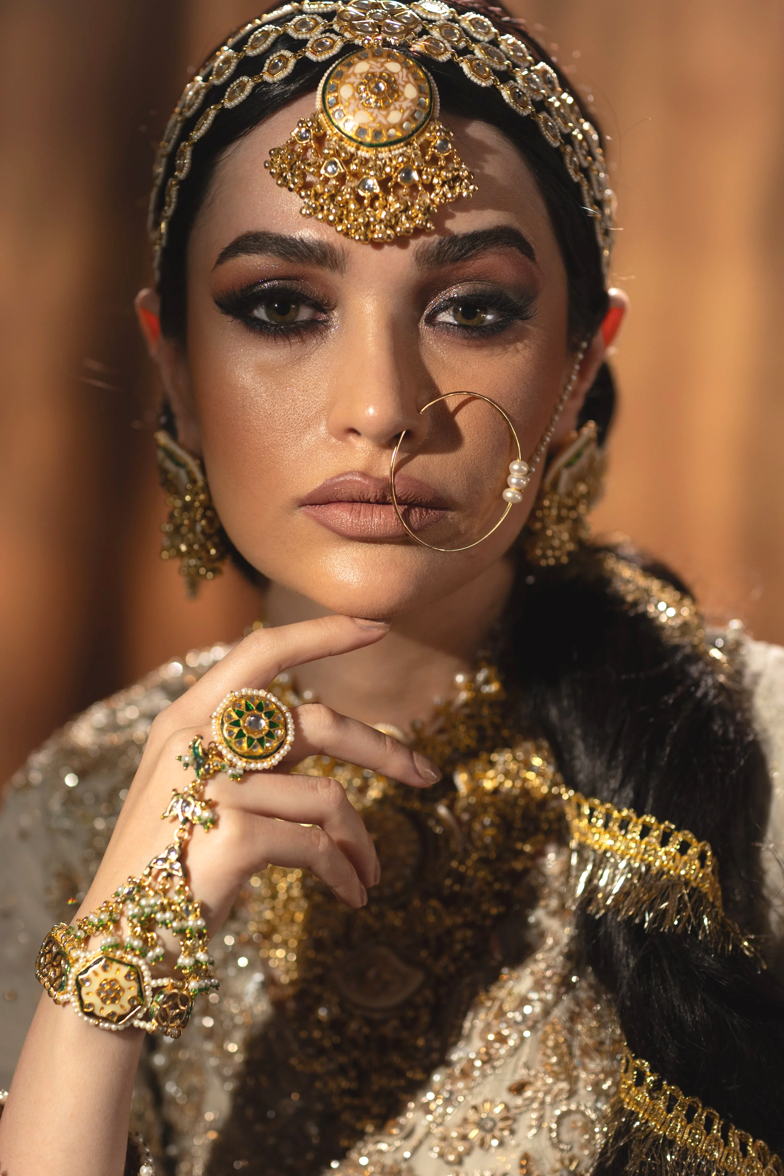 A woman wearing traditional Indian jewelry, including a gold headpiece, earrings, nose ring, and rings, with a braided hairstyle and elaborate embroidered outfit.