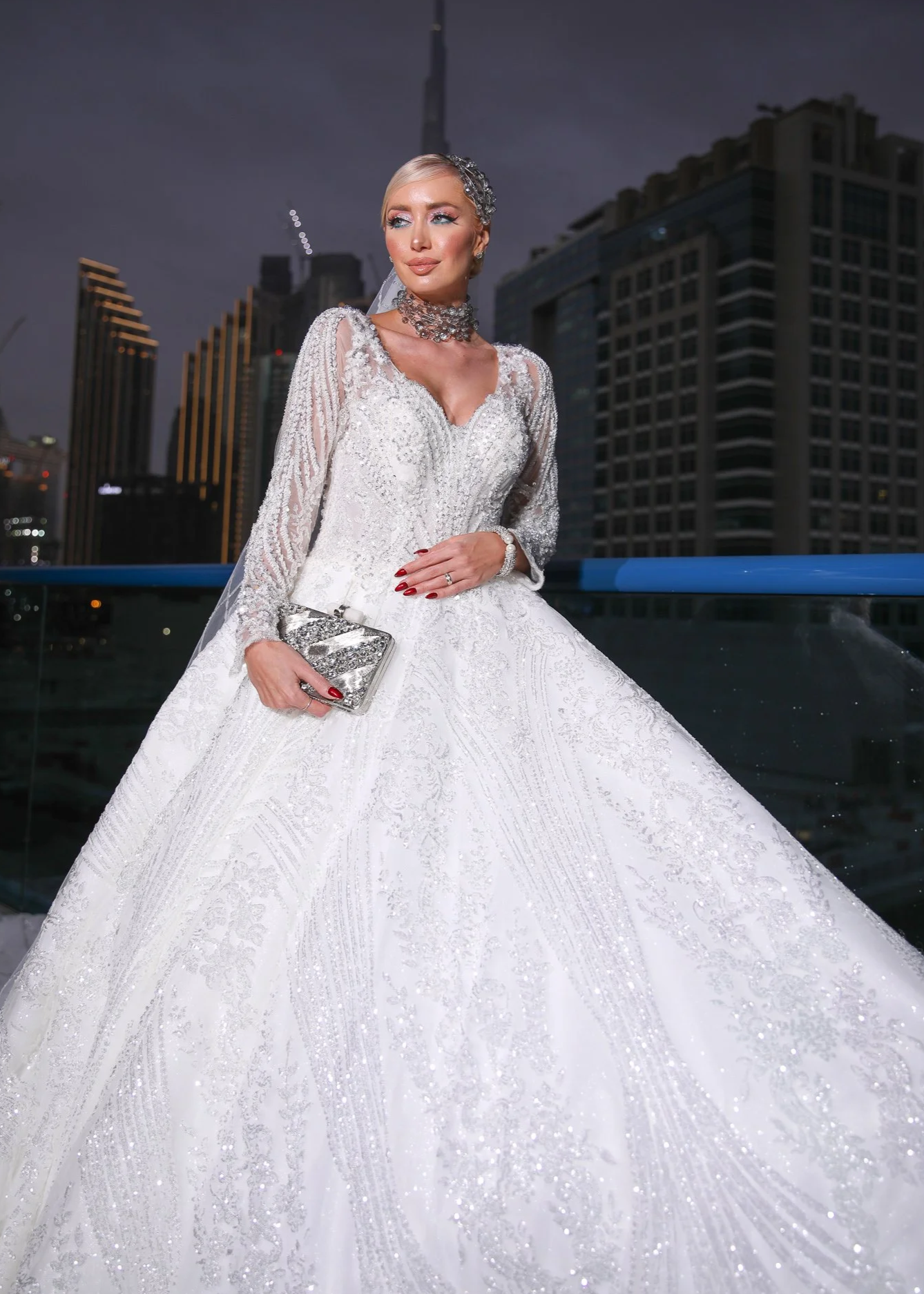 A woman in an ornate white wedding gown with lace and detailed embroidery, holding a small silver clutch, standing on a balcony with city buildings in the background at night.
