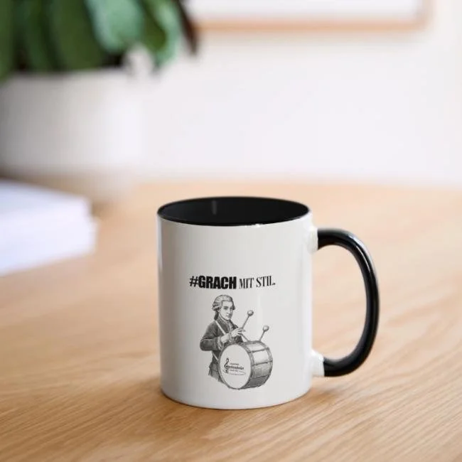 Weiße Tasse mit schwarzem Griff auf einem Holz Tisch, mit Aufdruck von einer Person mit Trommel und dem Text '#GRACH MIT STILL'