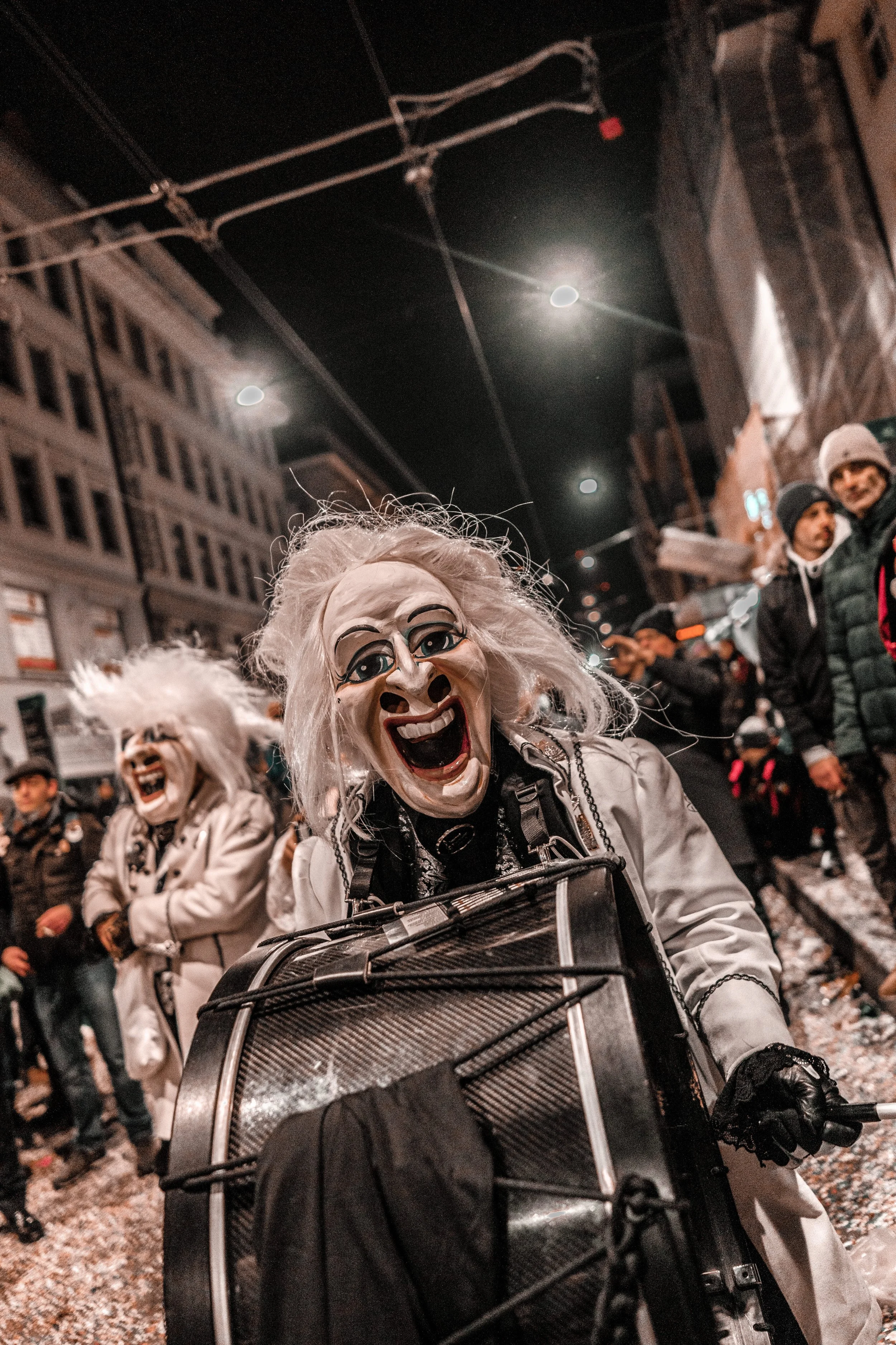 Menschen tragen bei einer nächtlichen Feier Masken mit lachenden Gesichtern und stimmen sich auf einen Karneval oder Fasnacht-Umzug in einer belebten Stadt ein.