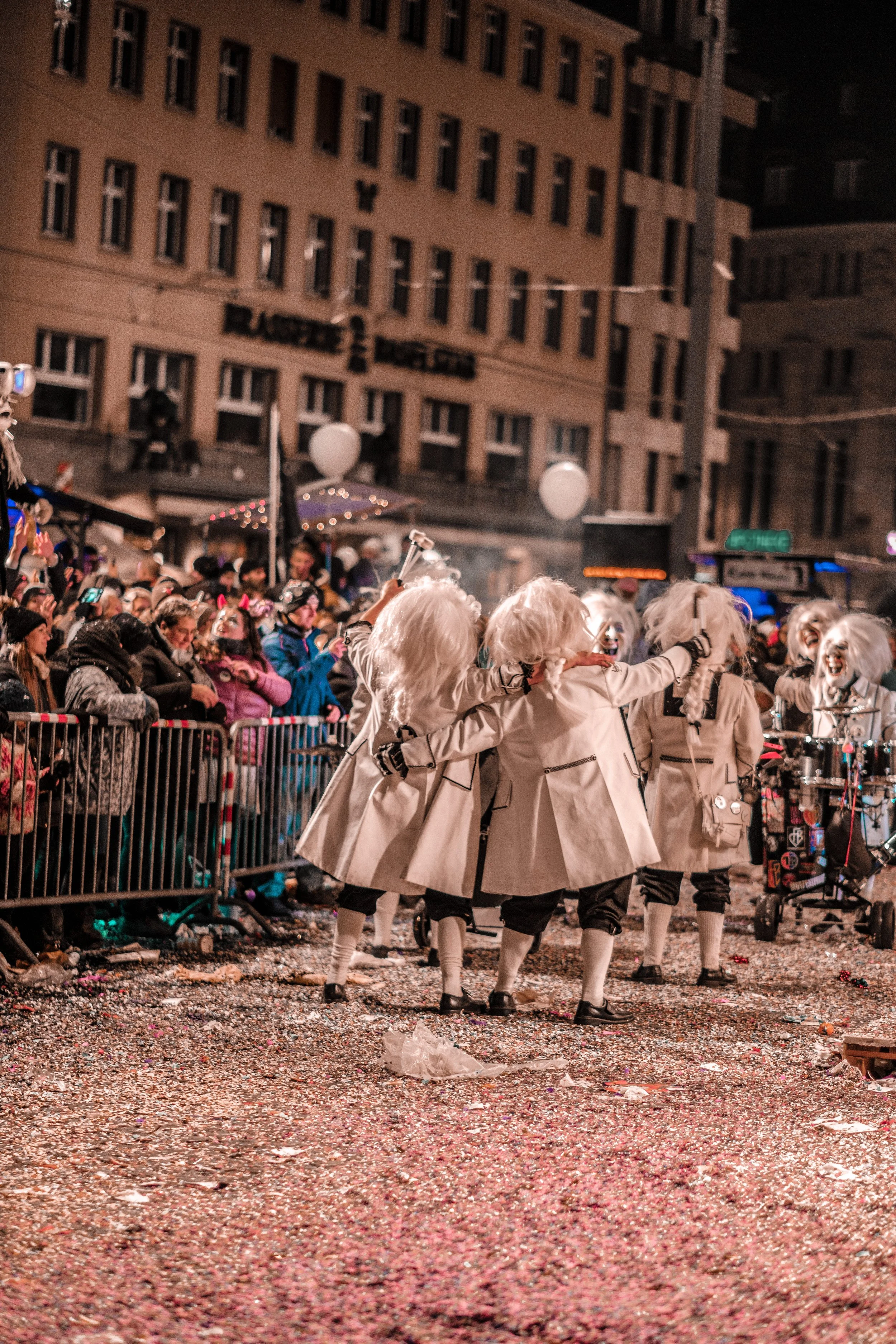 Gruppierung von Personen in Kostümen, die bei einem Straßenfest oder Umzug tanzen, mit einem Publikum im Hintergrund.