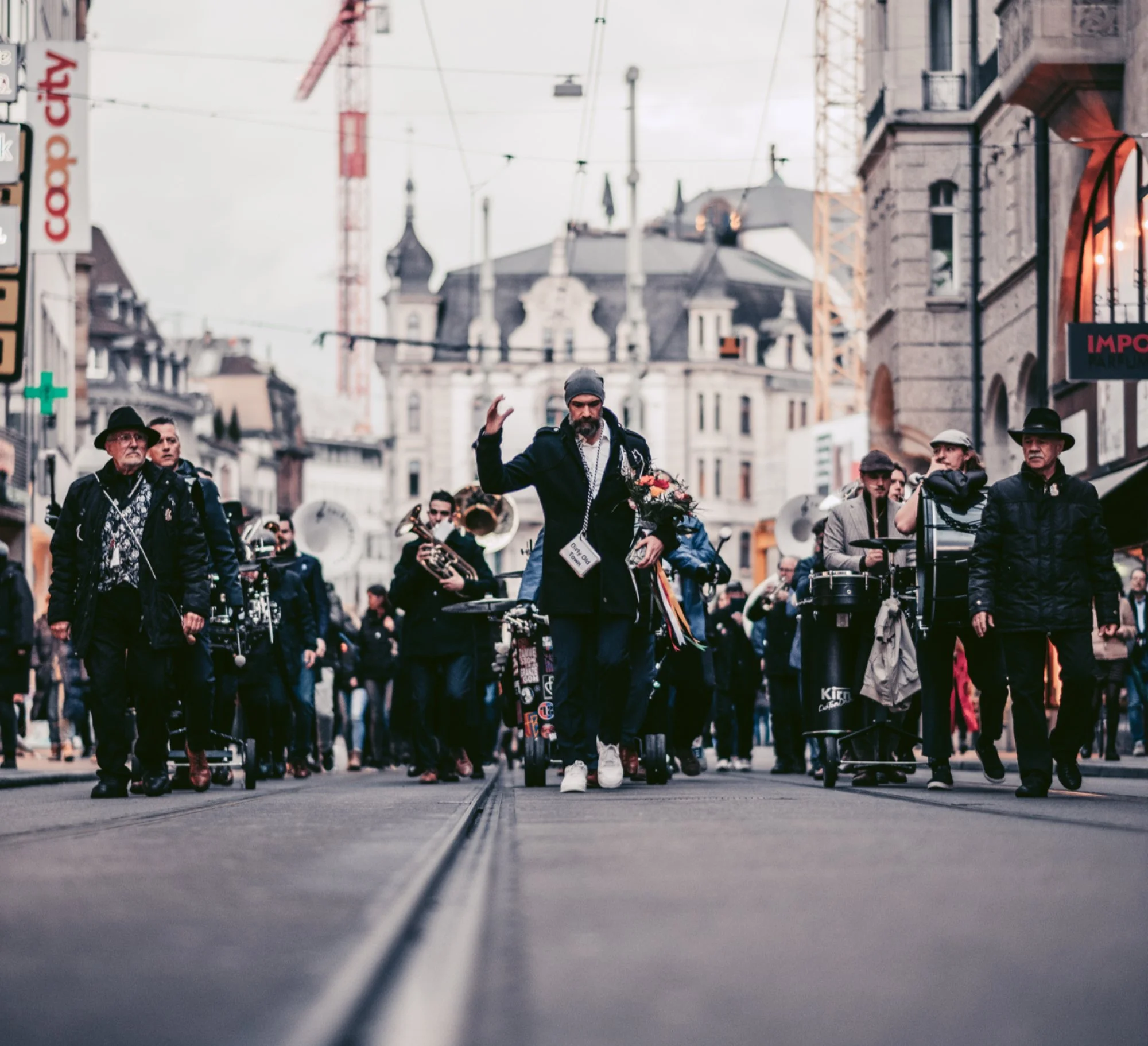 Menschen ziehen in einer Stadtstraße mit Straßenbahnschienen, einige tragen schwarze Hüte und Jacken, man sieht eine Gruppe von Musikern mit Instrumenten und eine Person in der Mitte, die eine Blume hält und winkt, davor auf dem Boden ist die Straße 