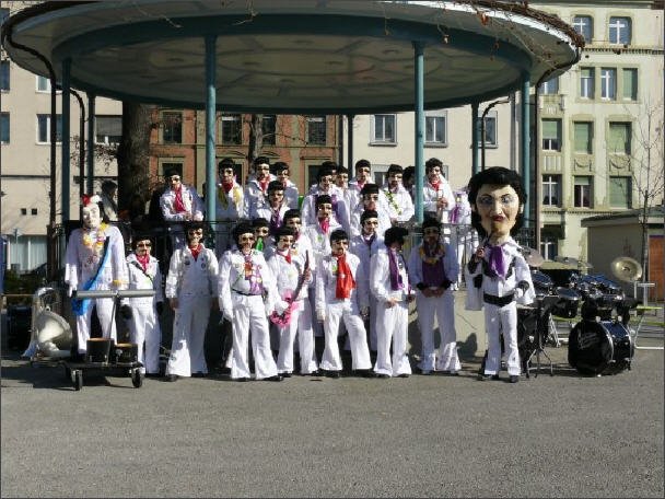 Gruppe von Menschen in weißen Anzügen, die vor einem Karussell stehen, mit einer großen Strohhutpuppe und Musikgeräten im Vordergrund, vermutlich auf einer Straße in einer Stadt.