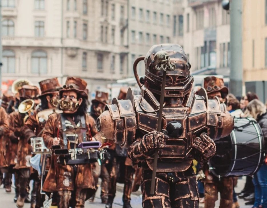 Menschen in Kostümen beim Karnevalsumzug, viele tragen Steampunk- oder Roboter-inspirierte Outfits, in einer Stadt mit historischen Gebäuden