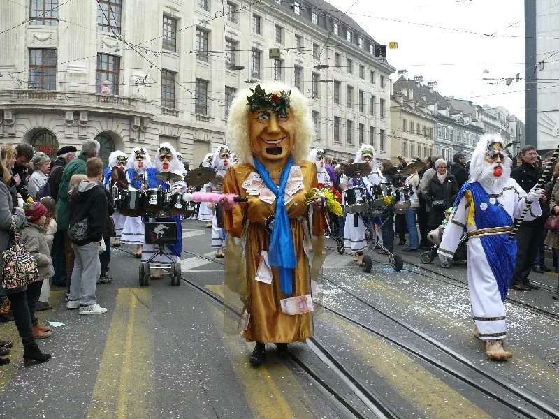 Bunte Karnevalsfiguren in Kostümen und Masken bei einem Straßenumzug in einer Stadt.