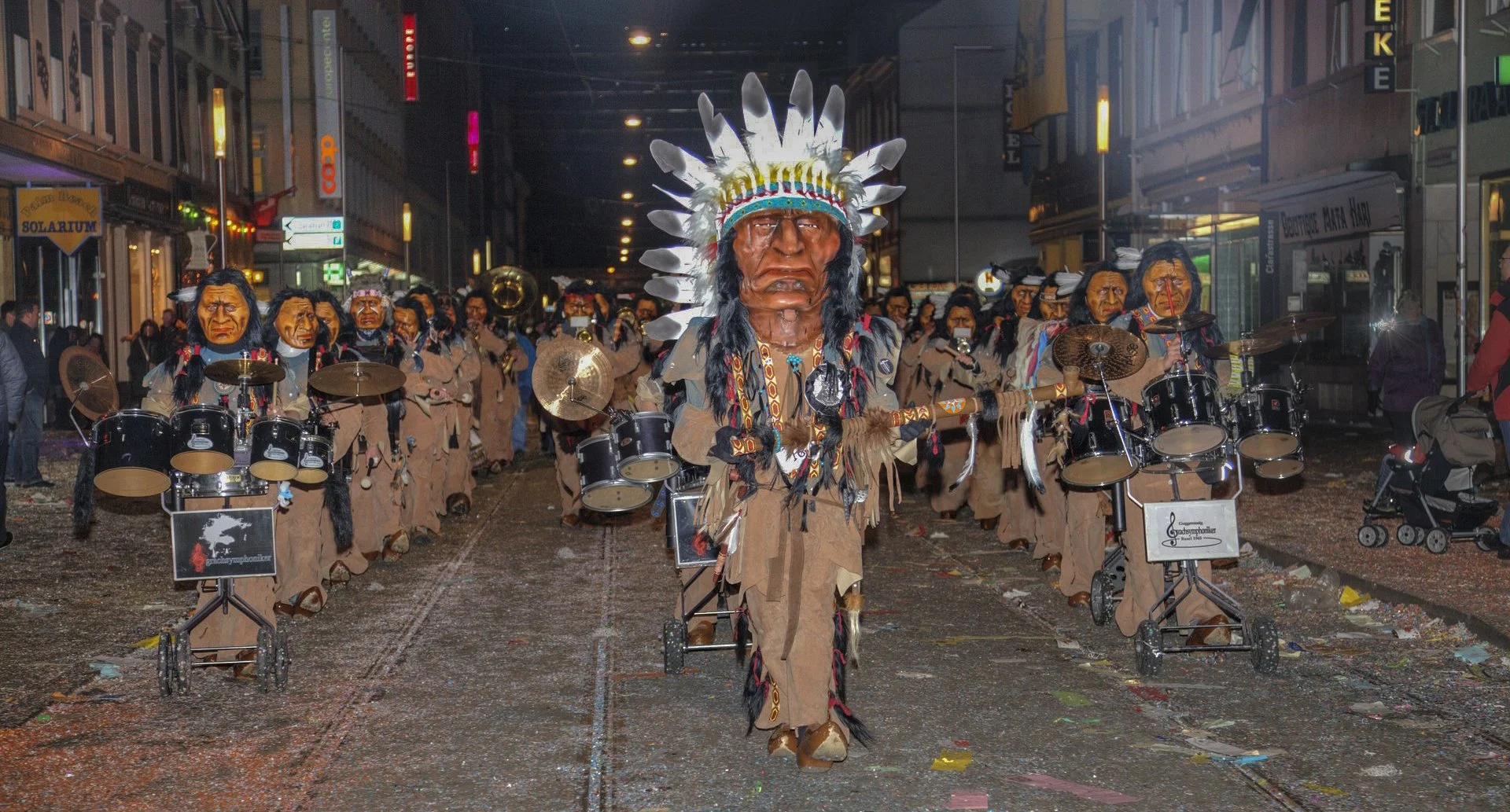 Eine Band mit Menschen in Indianer-Identitäten beim Rosenmontagsumzug in der Stadt, mit Kostümen, Masken und Musikinstrumenten auf der Straße bei Nacht.