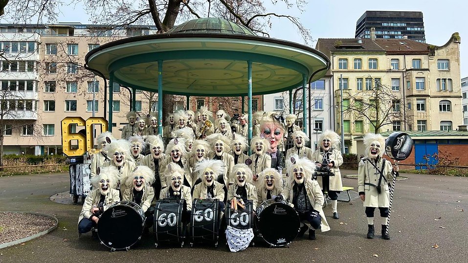 Gruppe verkleideter Menschen mit weißem, zotteligem Haar und weißen Masken, die in einem Park auf einer Holzbühne stehen, umgeben von Wohnhäusern, einige halten Musikinstrumente und eine große Maske in Form eines Gesichts.