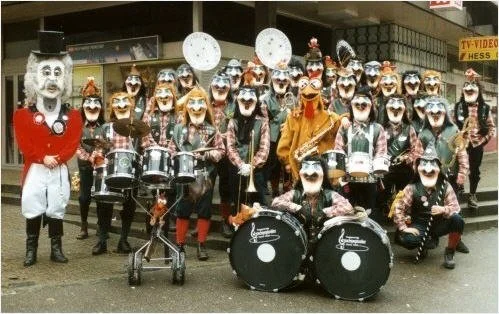 Eine Gruppe von Menschen, die in Karnevalskostümen mit clownartigen Masken, Musikinstrumenten und einem Quallen-Kostüm für eine Straßenparade oder Feier posieren.