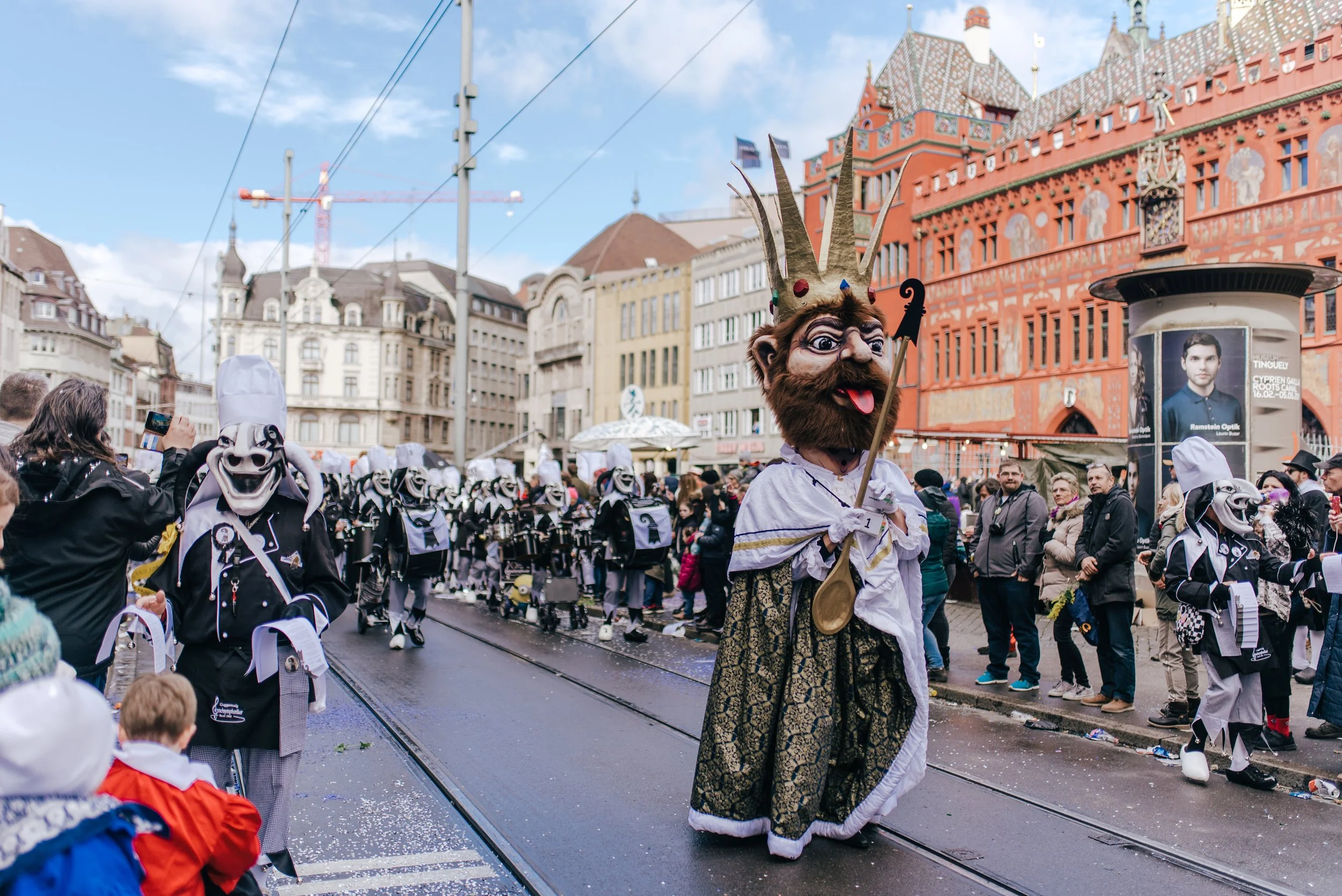 Menschen in bunten Kostümen und Masken, die an einer Parade auf der Straße teilnehmen, mit historischen Gebäuden im Hintergrund.