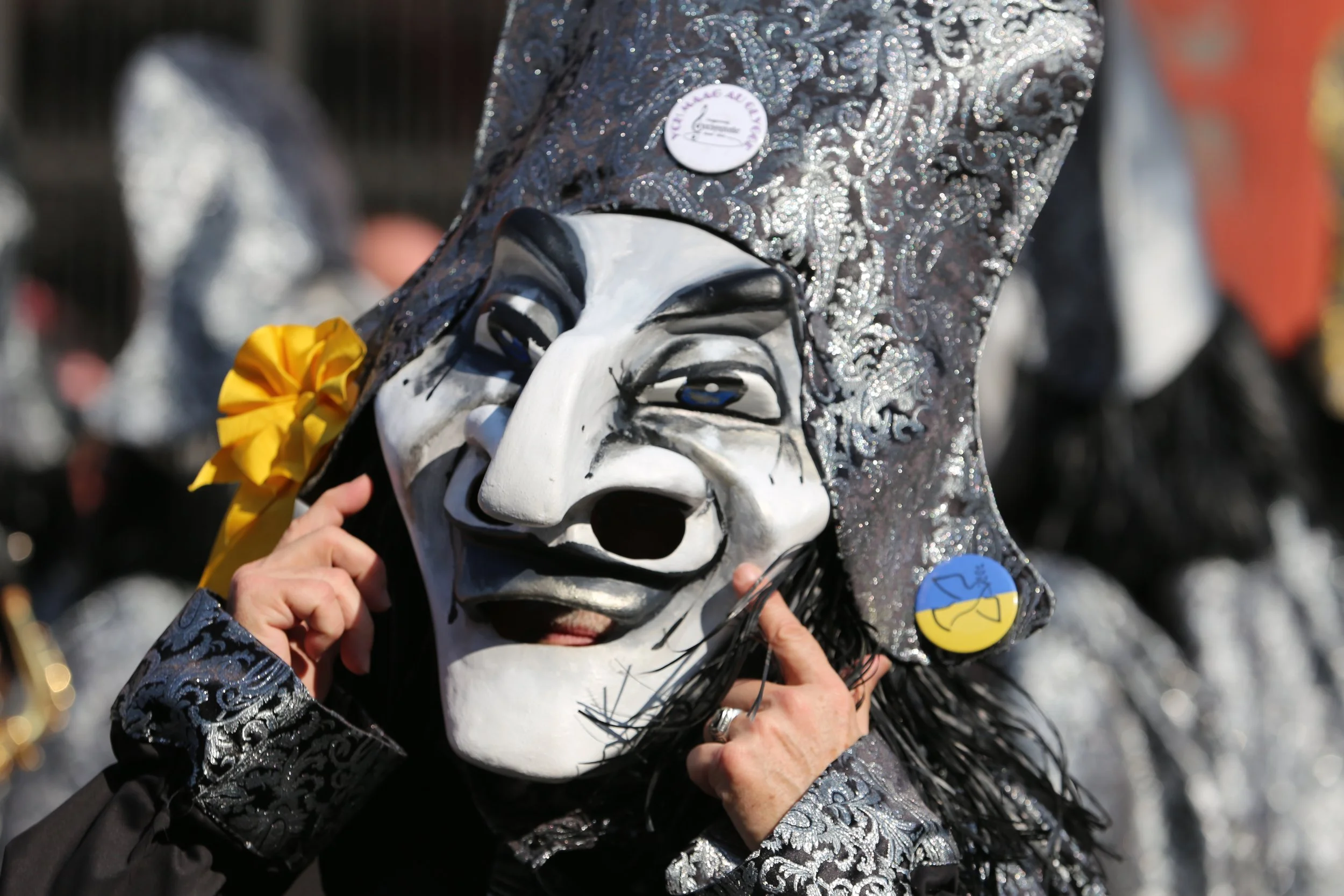 Eine Person trägt eine große, detaillierte Karneval- oder Festivalmaske mit einem lachenden Gesicht, weißen und schwarzen Farben und einem silber-glitzernden Kopfschmuck. Die Maske hat einen breiten Lachen-Ausdruck mit offenen Mund und hängenden Händ
