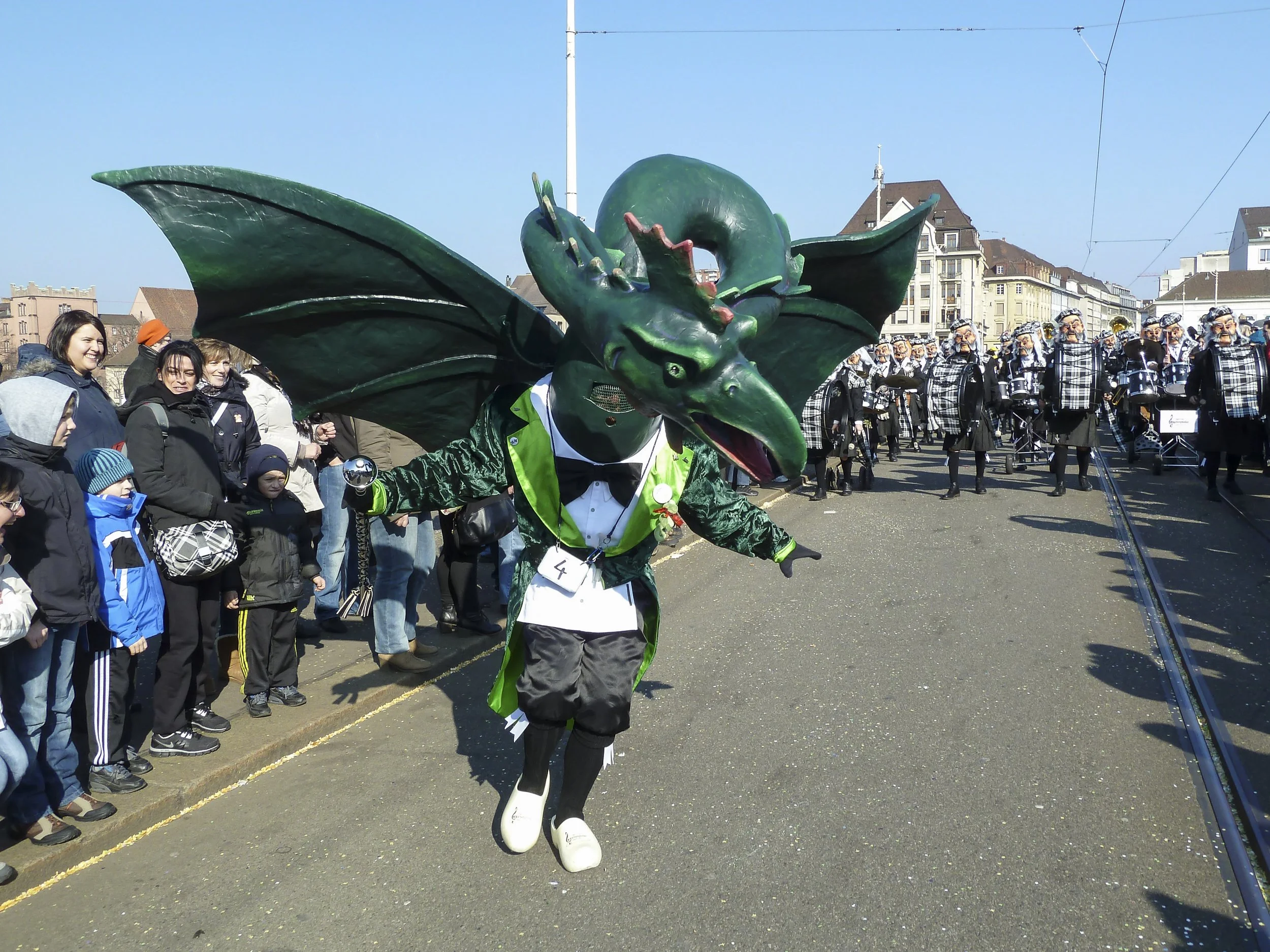 Mensch in Drachen-Kostüm bei einem Karnevals-Umzug, umgeben von Zuschauern und einer marschierenden Musikkapelle.