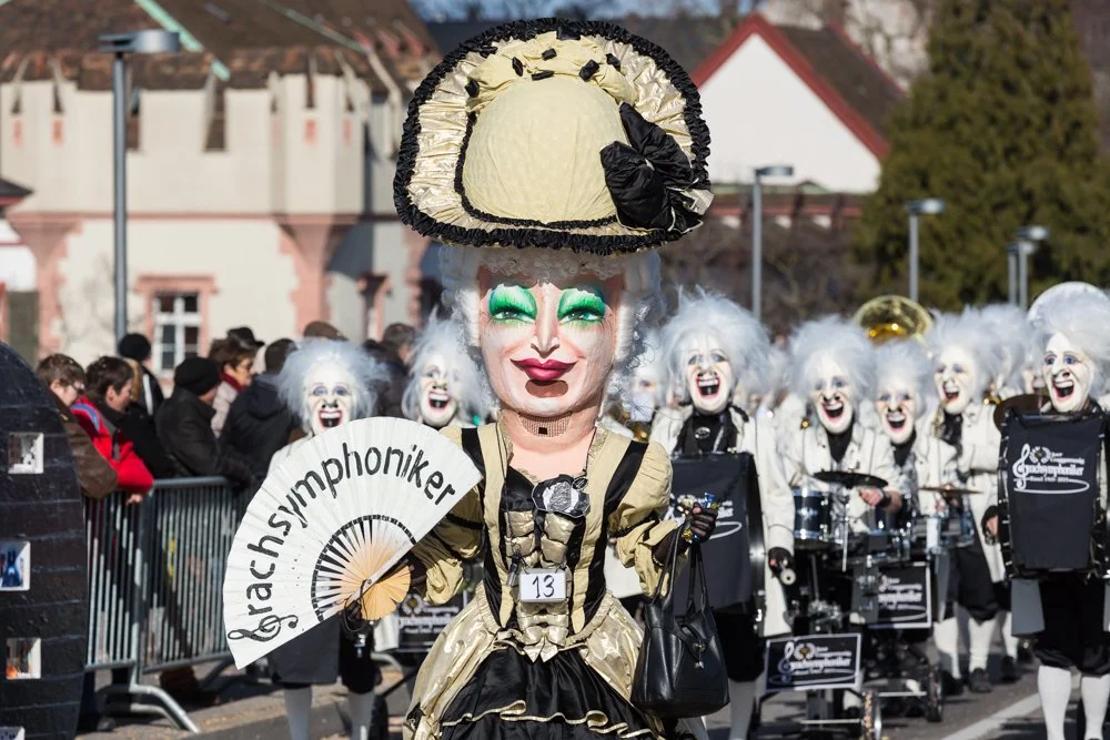 Eine Gruppe von Menschen in Karnevalskostümen, darunter eine Person im Vordergrund mit einem großen, eleganten Hut und einem geschnitzten Gesicht, die ein Fan mit der Aufschrift 'RachsyphoniKER' hält, während im Hintergrund Musikanten mit weißem Haar und schwarzen Masken auf einem Wagen spielen.
