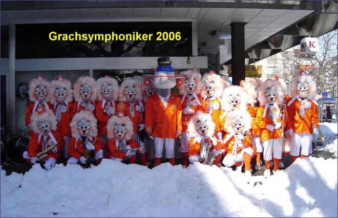 Gruppen von Menschen in festlichen Clown-Kostümen und Masken, die auf Schnee vor einem Gebäude mit einem Schild stehen, das 'Grachsymphoniker 2006' sagt.